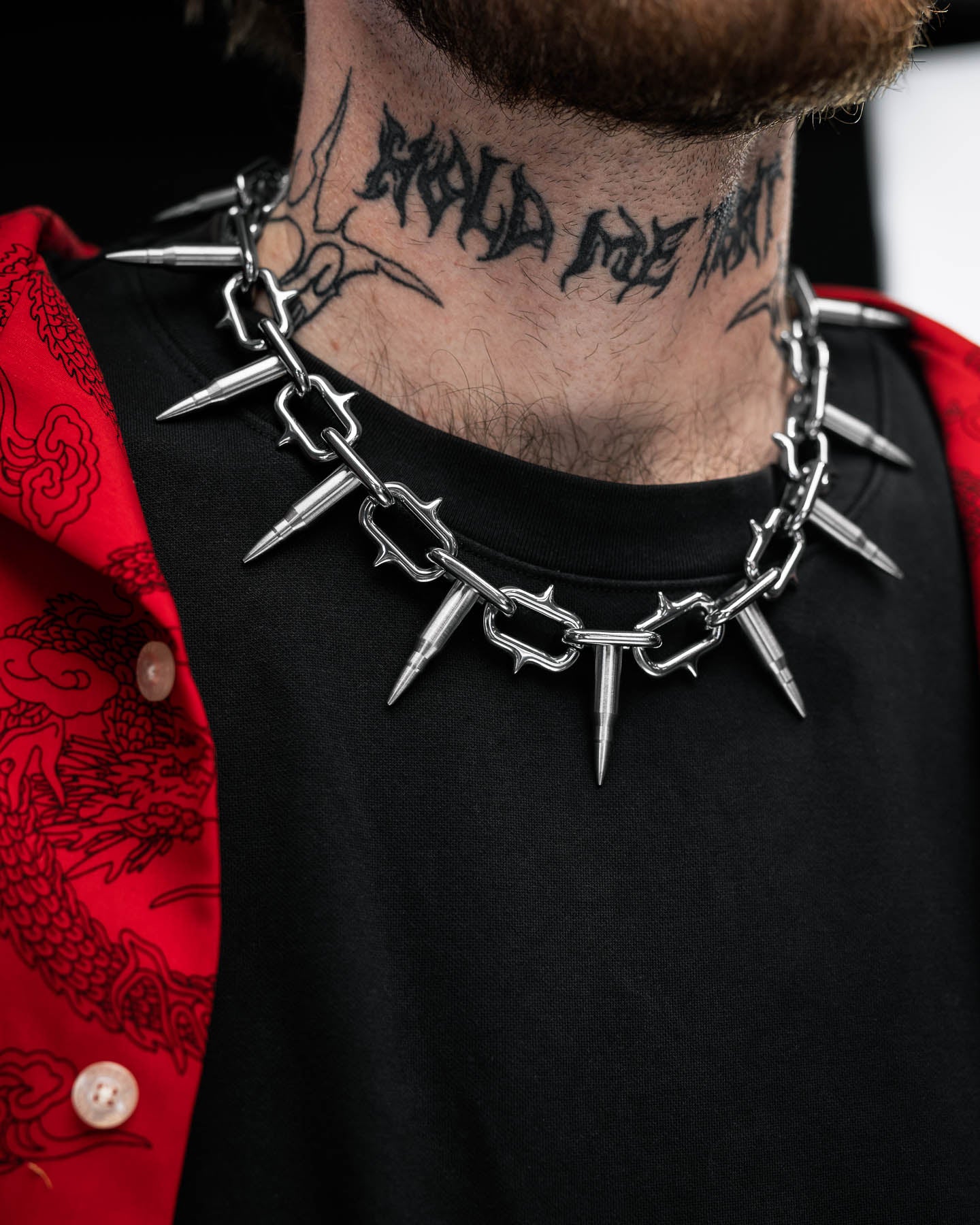 A person wearing a black shirt and red patterned jacket styled with the Statement Collective "Sweating Bullets" Necklace Chain—a chunky silver spiked piece. They have a neck tattoo with script and a design.
