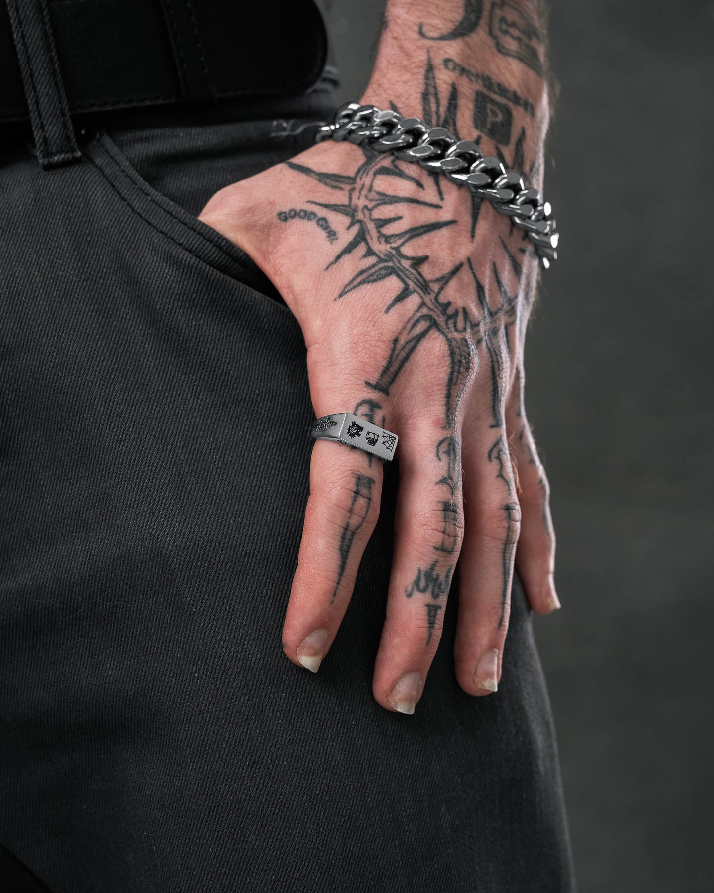 A tattooed hand in black jeans rests on a pocket, wearing a chunky chain bracelet and the Tattoo Signet Ring—a stainless steel ring featuring intricate symbols—set against a dark background.