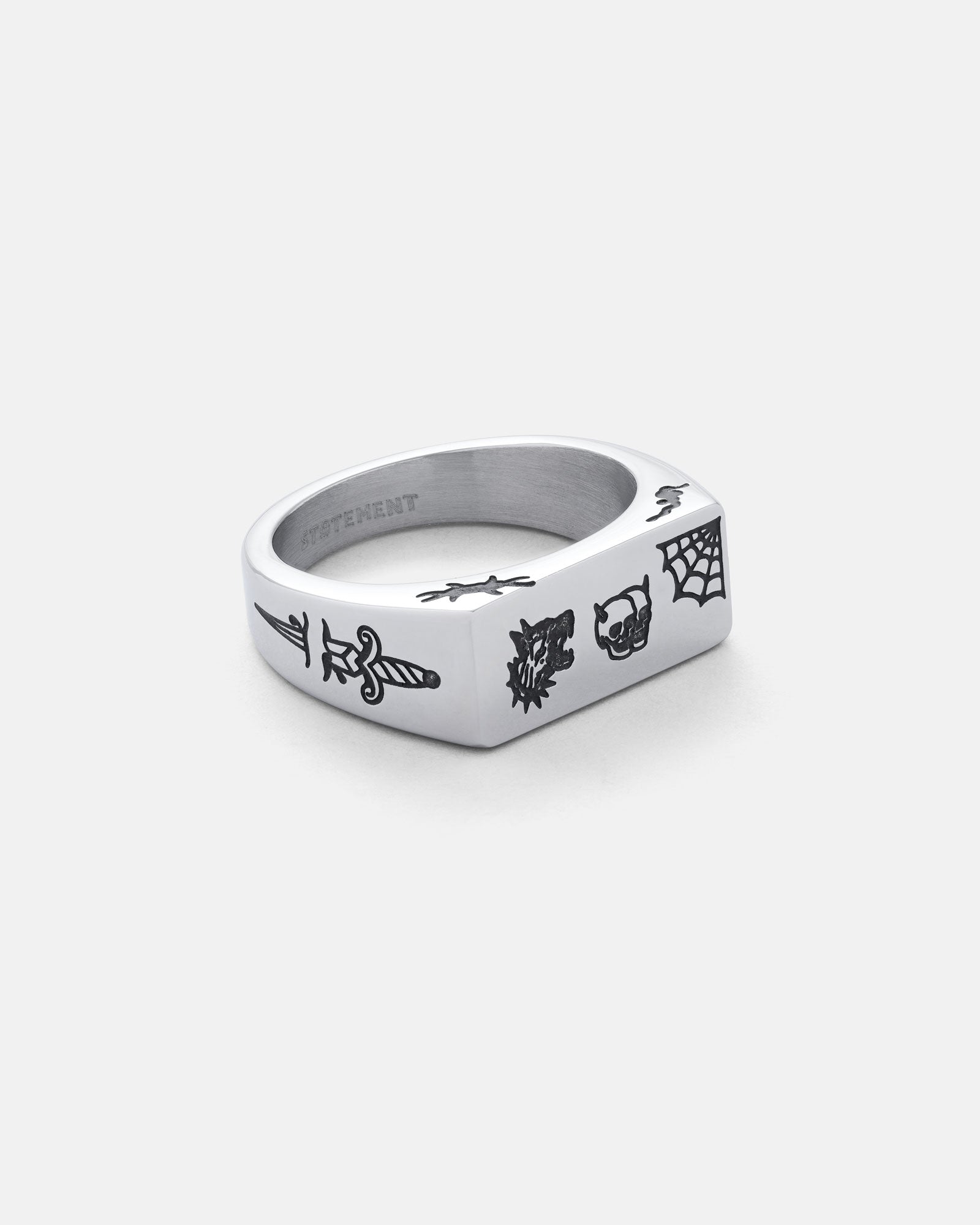 The Tattoo Signet Ring is a stainless steel ring with a flat rectangular face, engraved with sword, dragon, skull, and spiderweb tattoo motifs, shown on a plain white background.