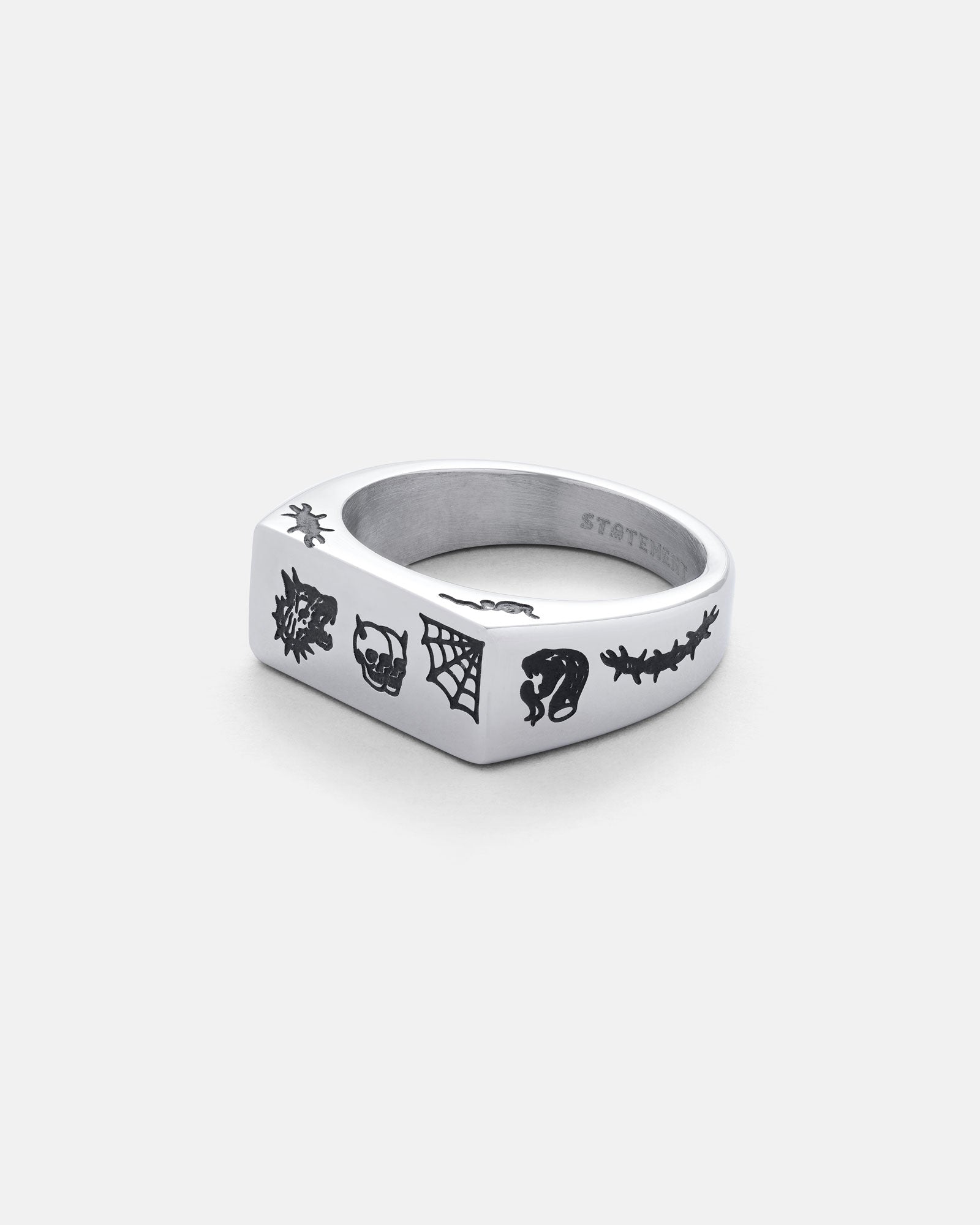 The Tattoo Signet Ring is a stainless steel ring with a flat edge, engraved with tattoo-inspired designs—spider, web, skull, face, and thorny branch—shown against a plain white background.