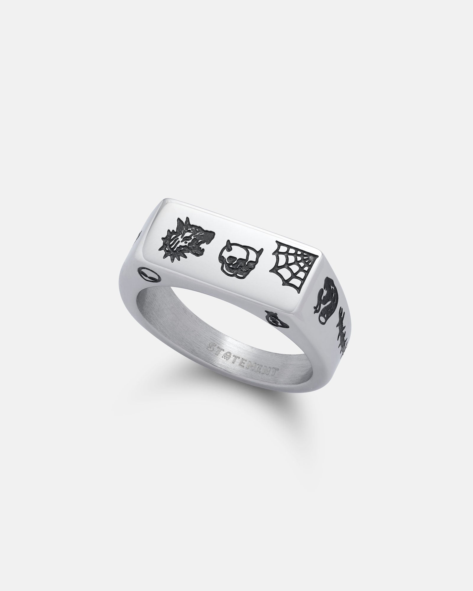 The Tattoo Signet Ring is crafted from 316L stainless steel and features a flat silver top engraved with bold black tattoo flash art—dragon, skull, spider web, lightning bolt, and eye—on a white background.