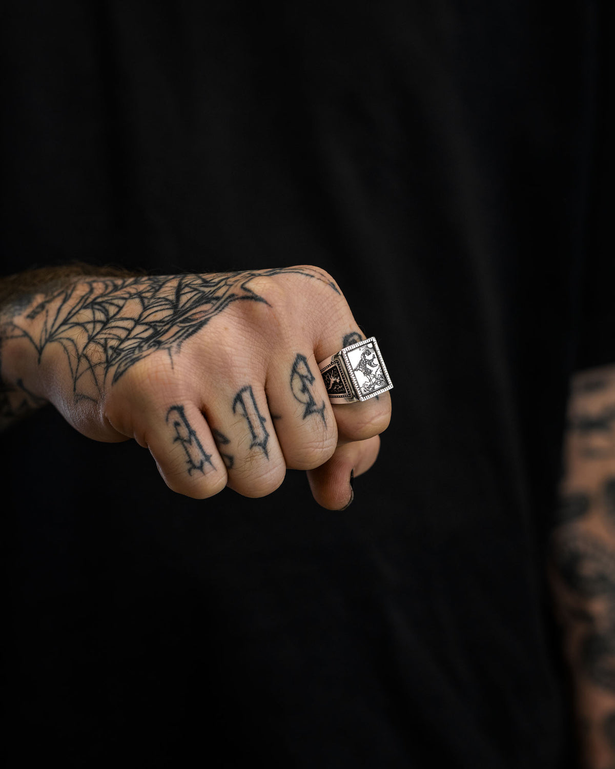 A tattooed hand with a spiderweb tattoo and the letters A L I E across the fingers wears the Statement Collective “Try Hard Fool” ring, a large silver piece featuring a detailed engraved image. Set against a black background.