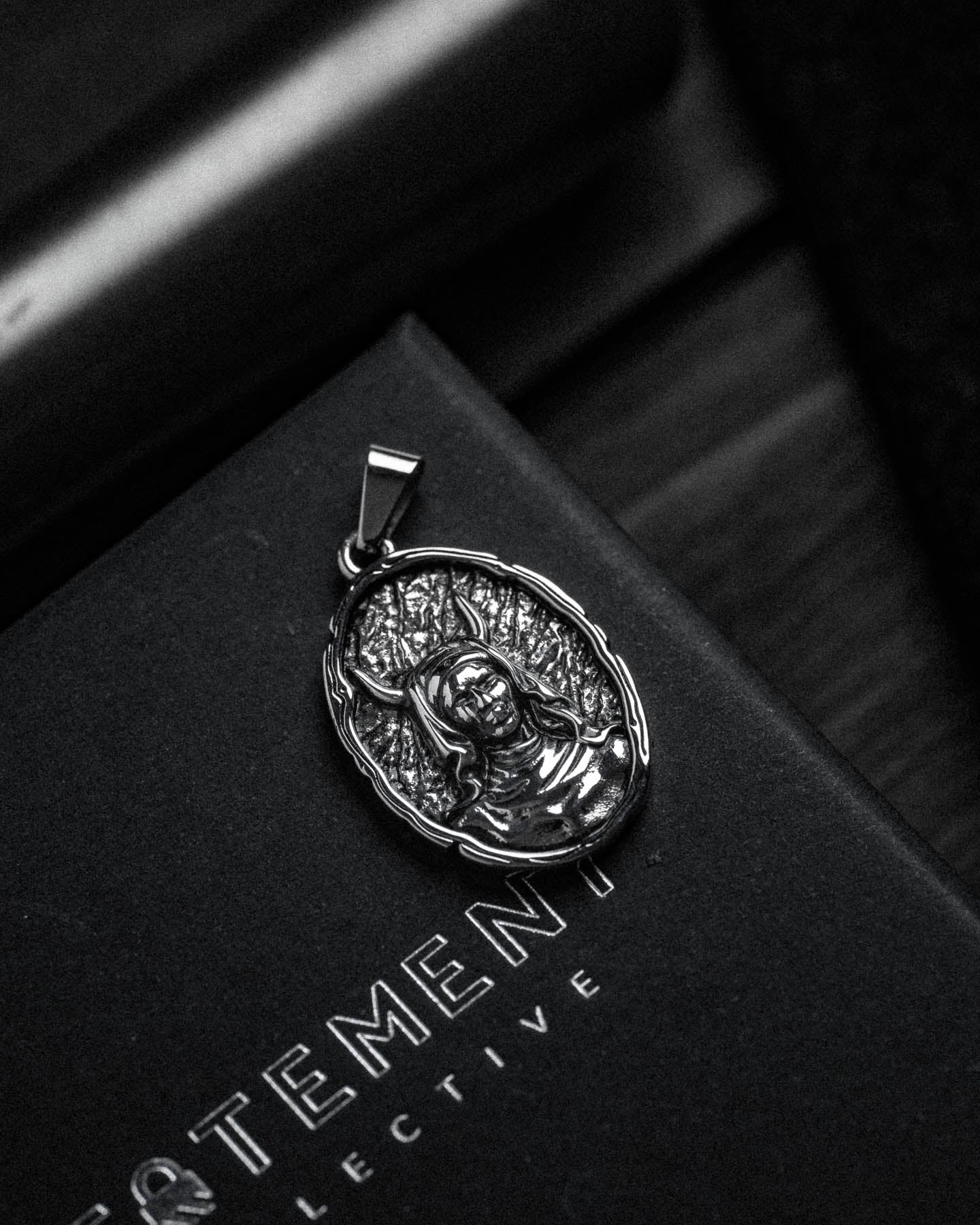 The Unholy Pendant, a silver gothic jewelry piece depicting a raised figure with a tree-like backdrop, rests on a black box showing partially visible white text: TEMENT and LECTIVE.