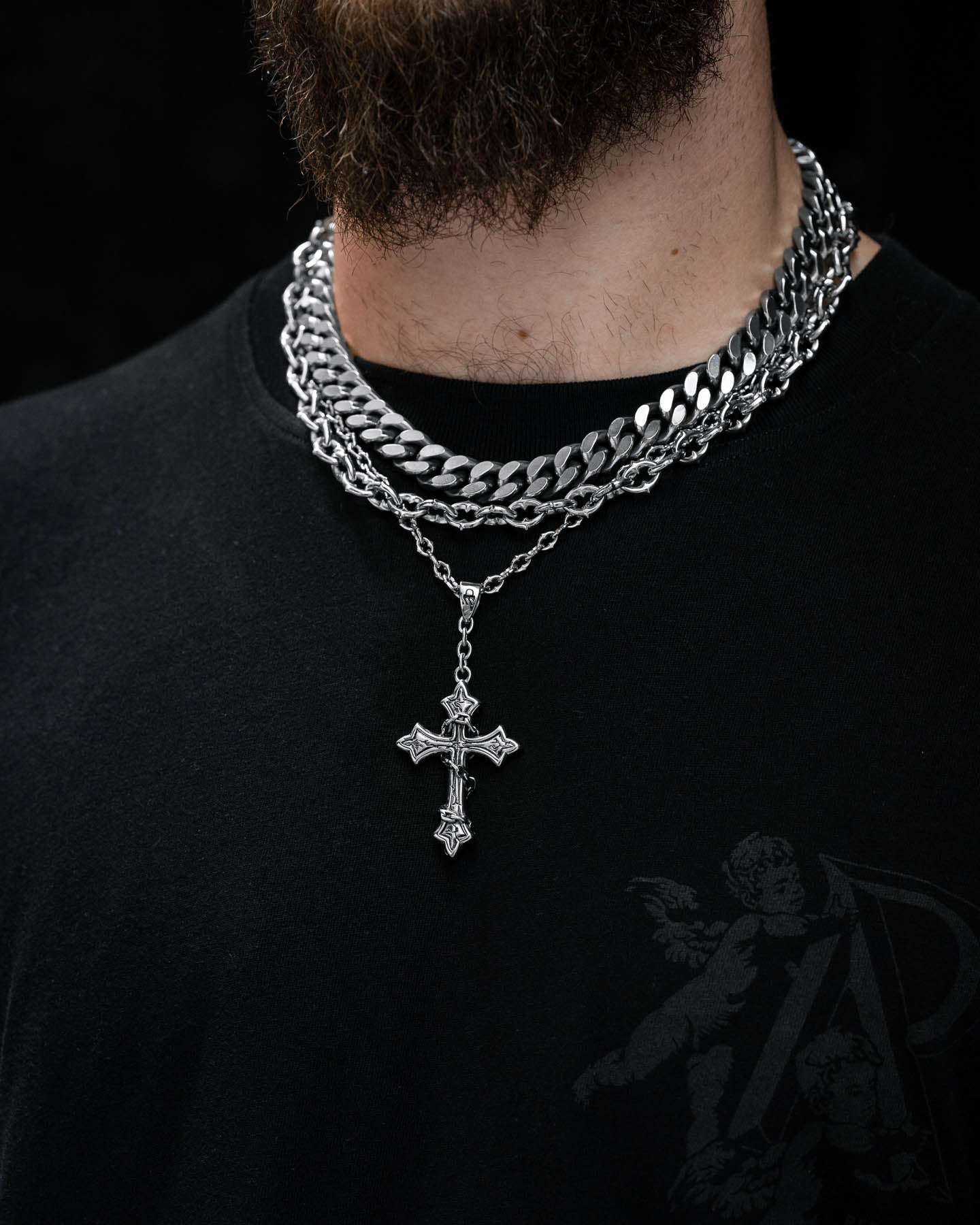 Wearing the Untouchable Stack, which includes a chunky Cuban chain and a Ruins Cross Pendant, this person pairs their black shirt’s decorative design with bold silver chains. The dark background highlights only their lower face and neck.