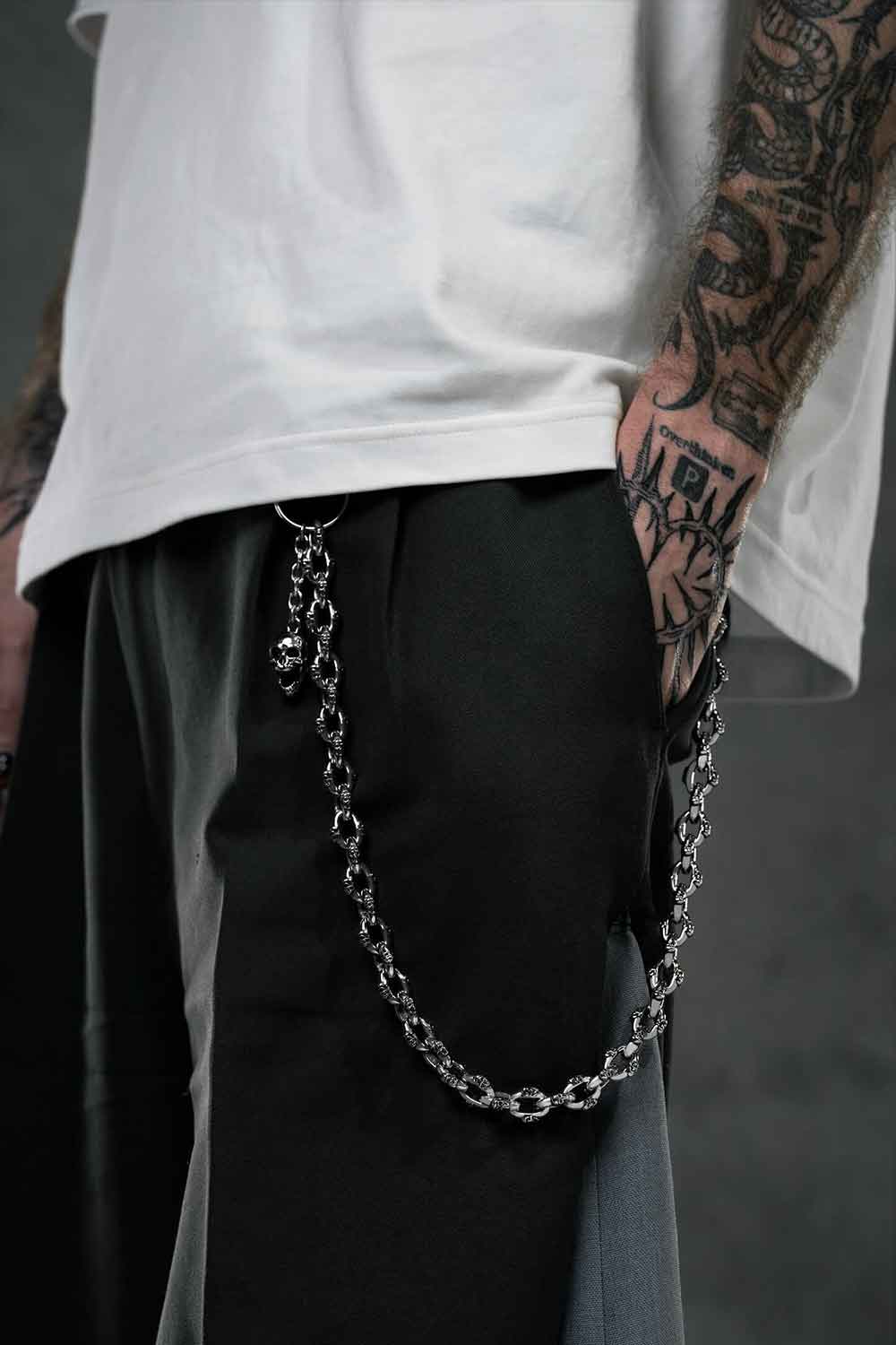 A person wearing black and gray pants with the "Boneyards" Skull Wallet Chain clipped to the pocket stands with a tattooed hand in the pocket, a partially visible white shirt, and a dark, blurred cement wall background.