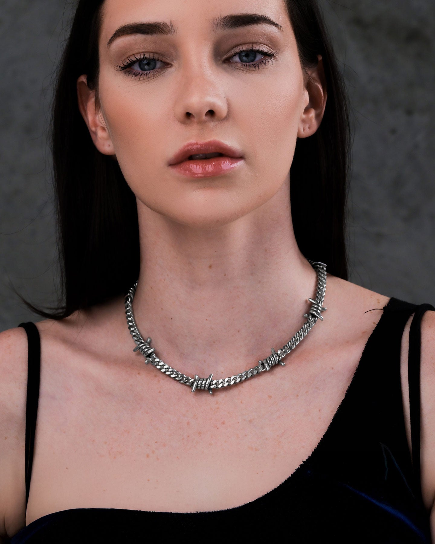 A woman with straight dark hair and light skin wears a black top and the 6mm Barbed Wire Cuban Chain necklace, standing against a dark, textured background.