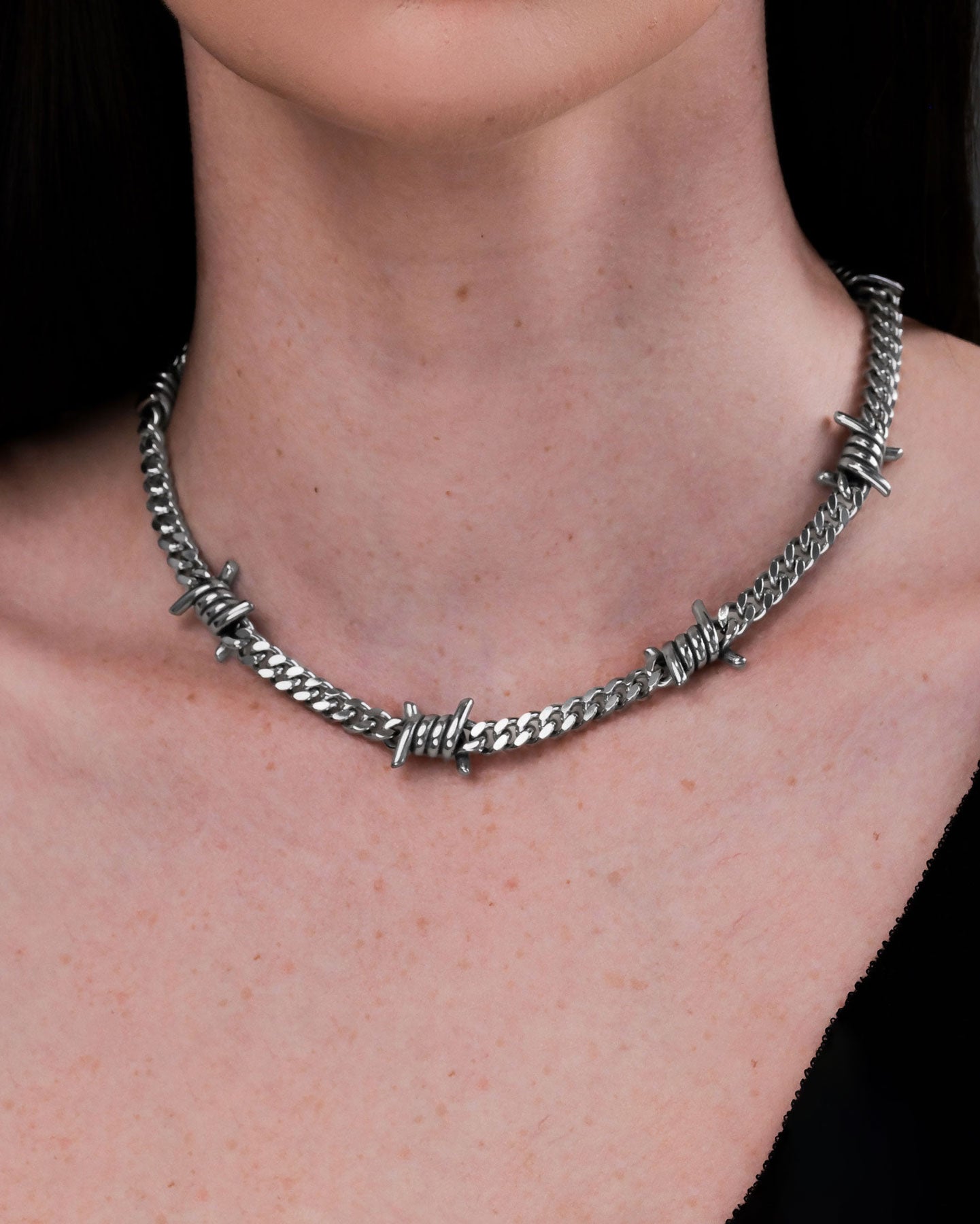 A close-up of a person wearing the 6mm Barbed Wire Cuban Chain, a bold and durable silver necklace crafted to resemble barbed wire, resting elegantly on their neck and collarbone and paired with a black top. This piece is waterproof and non-tarnish.