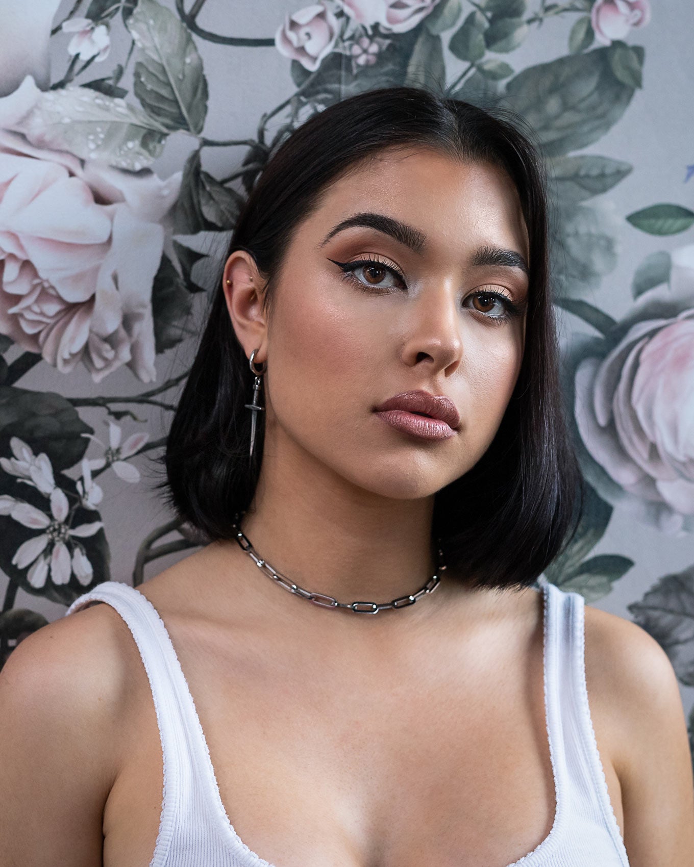 A woman with straight dark hair and winged eyeliner wears a white tank top, a Statement Collective Paperclip Chain necklace, and a cross earring, standing before floral wallpaper with large pink and white roses.