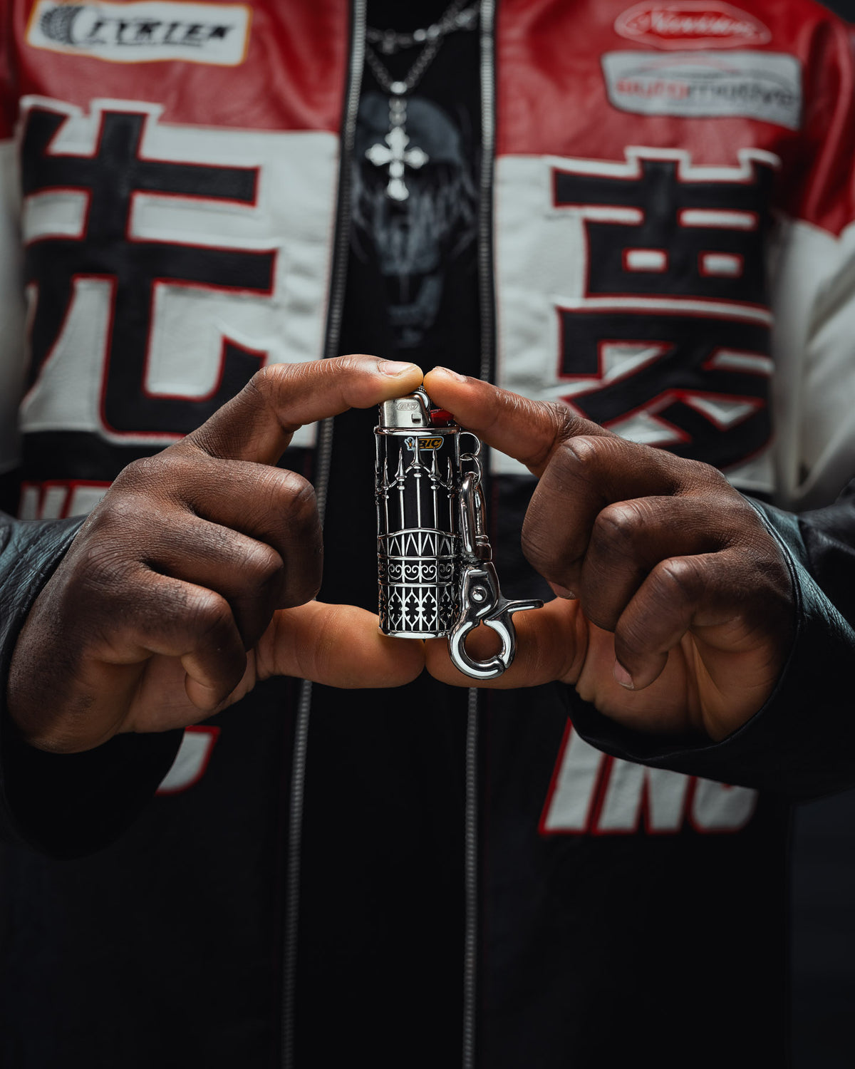 A person wearing a black and red jacket holds up the Statement Collective “Wrought Iron” Lighter Sleeve with both hands, showcasing its detailed black and white design on a sleek stainless steel case.