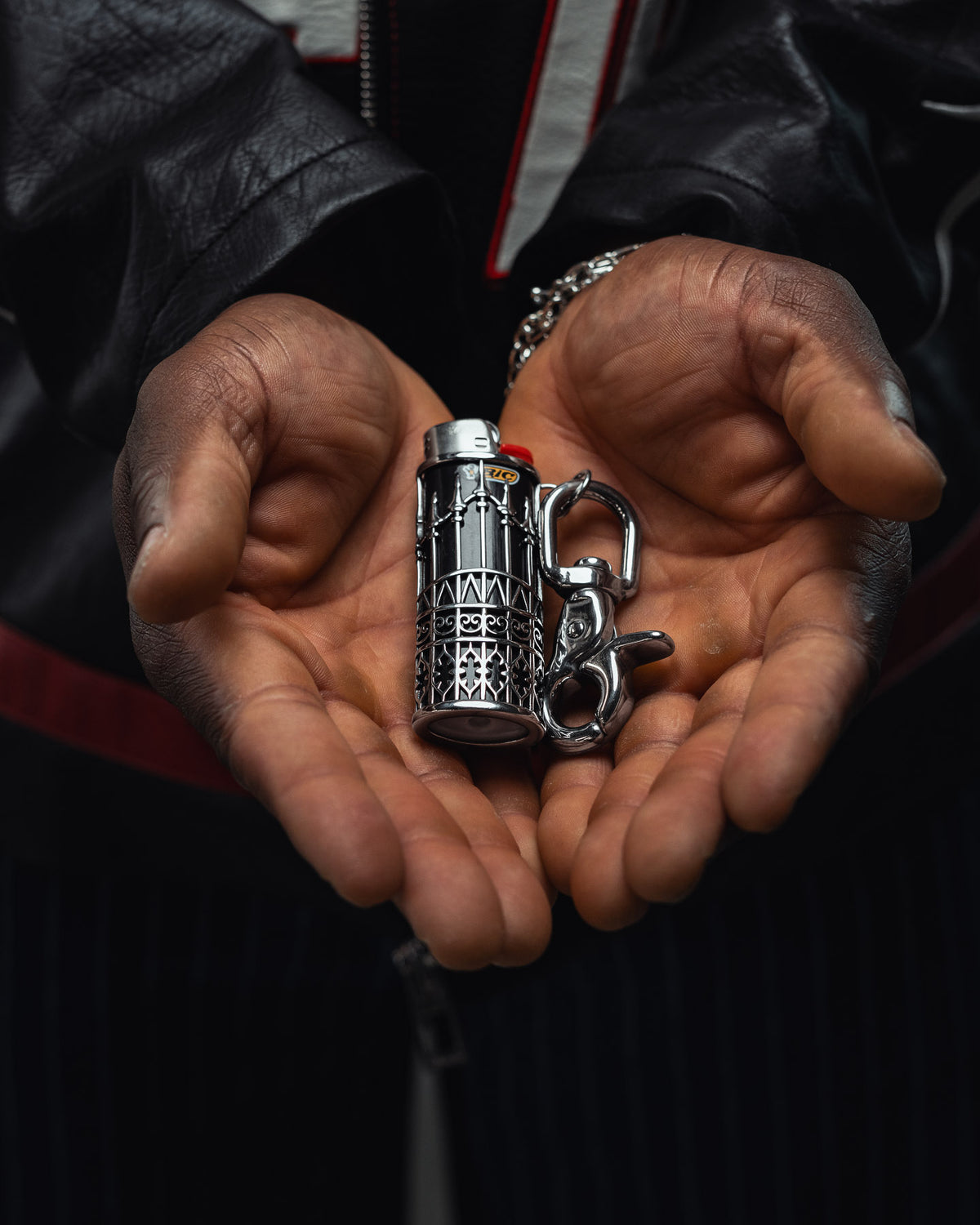 A person wearing a black leather jacket displays the Statement Collective “Wrought Iron” Lighter Sleeve—a detailed, ornate stainless steel lighter case and keychain—in their open palms, with a chain bracelet visible on their wrist.