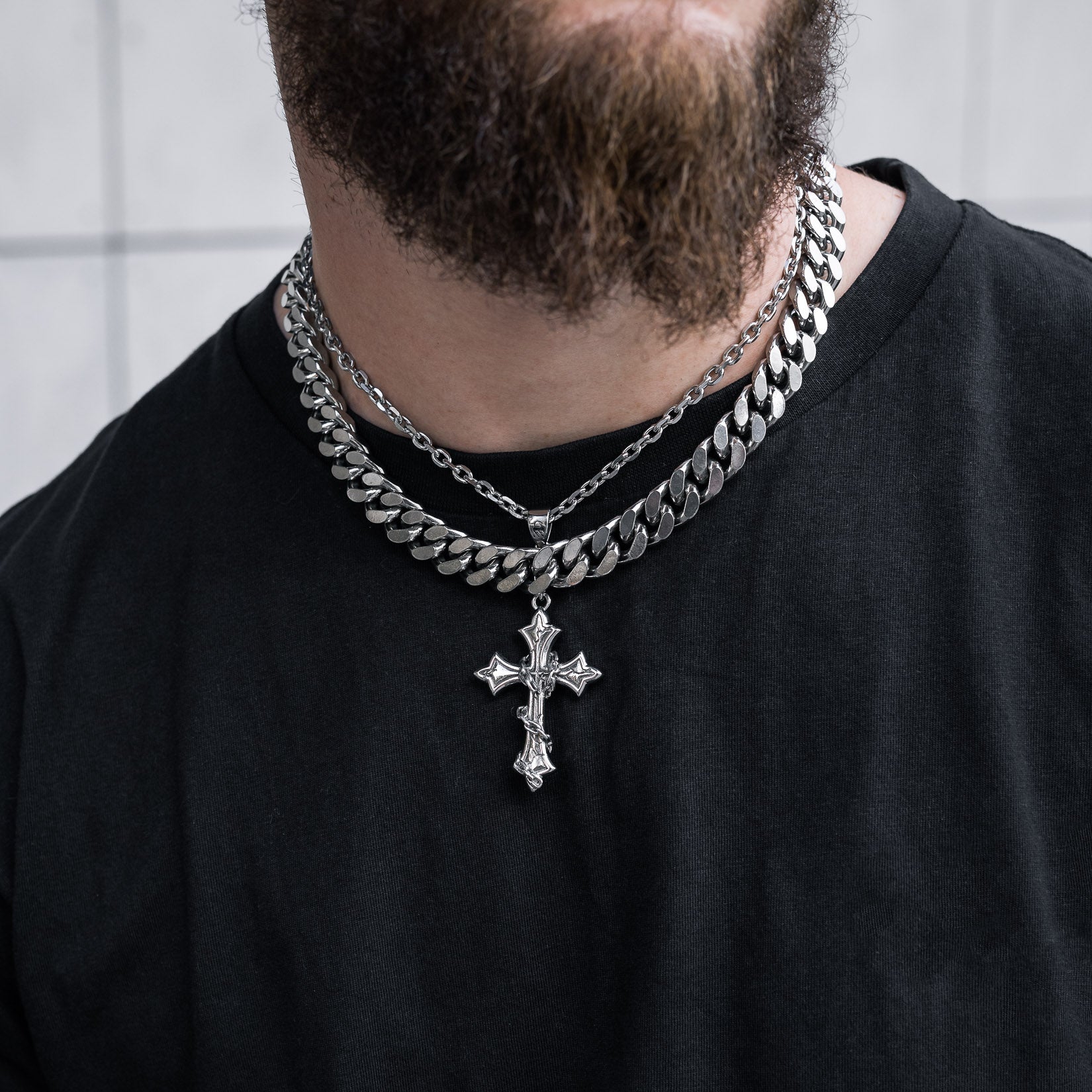 A person wears a black shirt with layered silver chains, including a 13mm Chunky Cuban Chain and an ornate cross pendant. The image is cropped to the neck and upper chest.