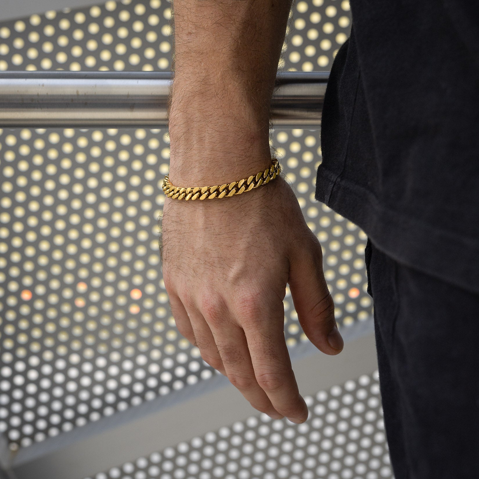 A close-up shows a person's left hand wearing the 9mm Cuban Bracelet (Gold), set against a metallic, perforated background, with the individual dressed in black.