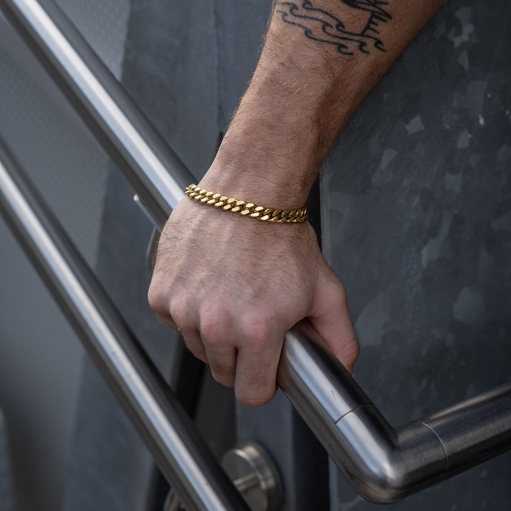 A tattooed hand grips a metal railing while wearing the 9mm Cuban Bracelet (Gold) near a textured metal wall.