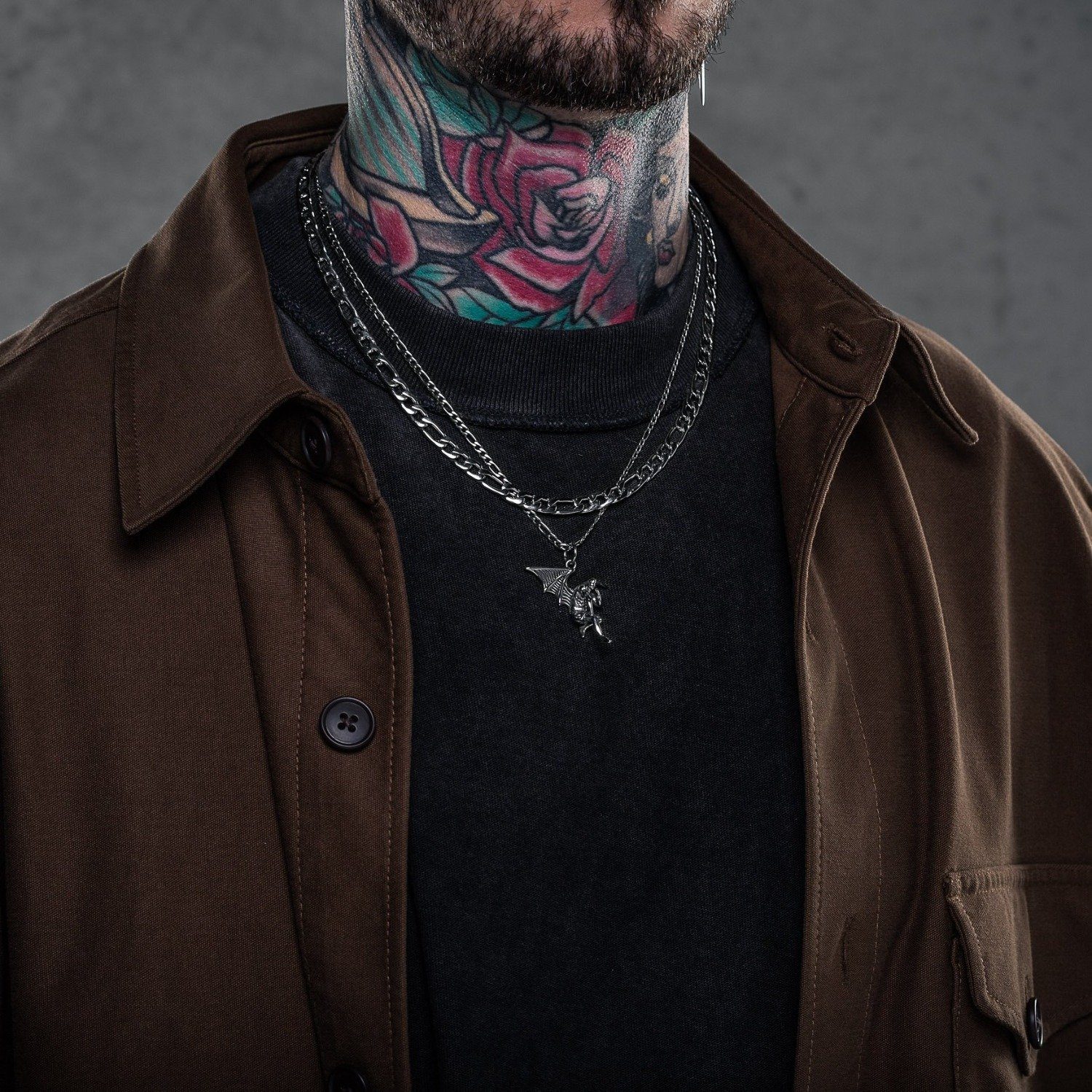 man with black shirt and brown jacket wearing angel necklace in set against concrete background