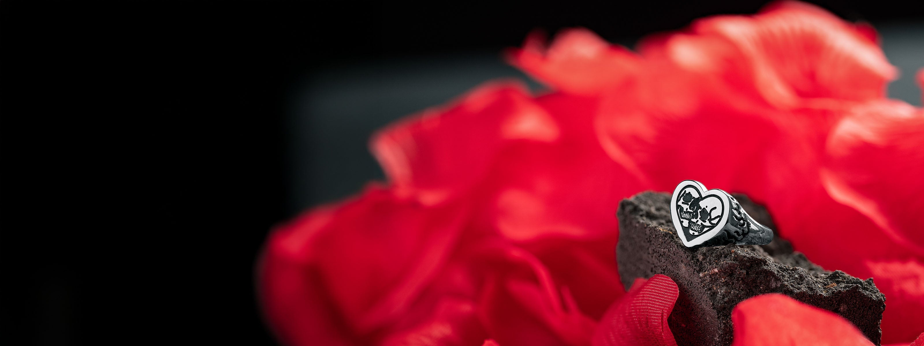 A heart-shaped ring with a black and white design rests on a piece of dark stone, surrounded by vibrant red rose petals, against a dark blurred background.