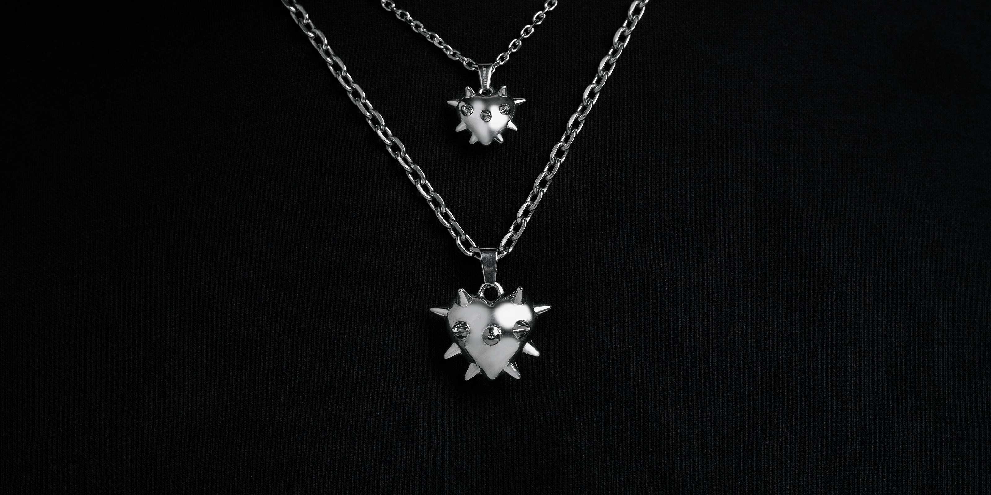 Two silver chain necklaces with spiked heart pendants are displayed against a black background, with one necklace shorter and the other longer.