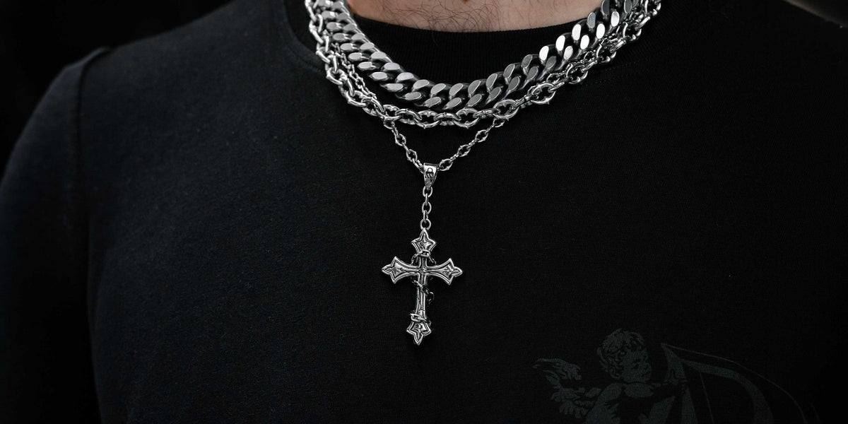 A man with a beard wears a black shirt, a white sherpa jacket, and two gold chains, one with a sword pendant and the other a thick curb link chain.
