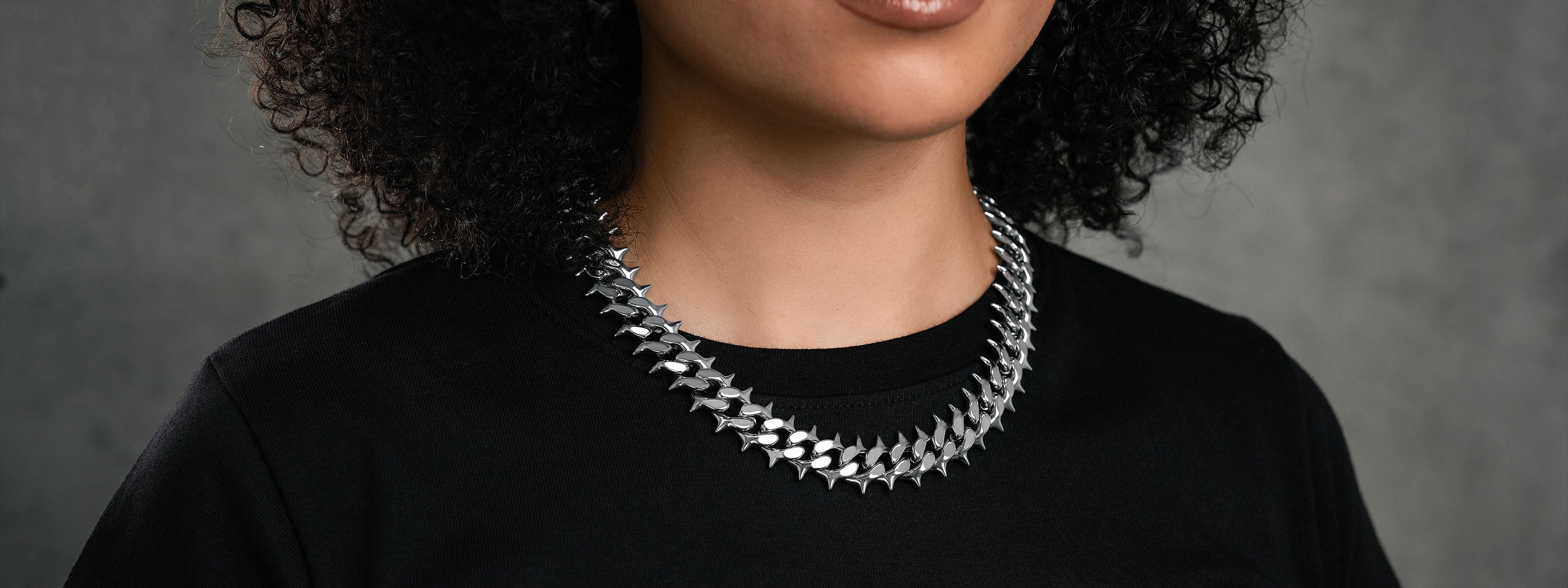Close-up of a woman with curly hair wearing a black shirt and a bold, silver, spiked necklace against a gray background. Her face is partially visible, showing only her lips and chin.