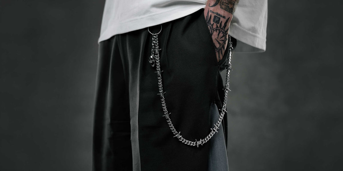 A person wearing black pants with a silver chain, white shirt, and visible tattoos on their hand and arm, standing against a dark, blurred background.