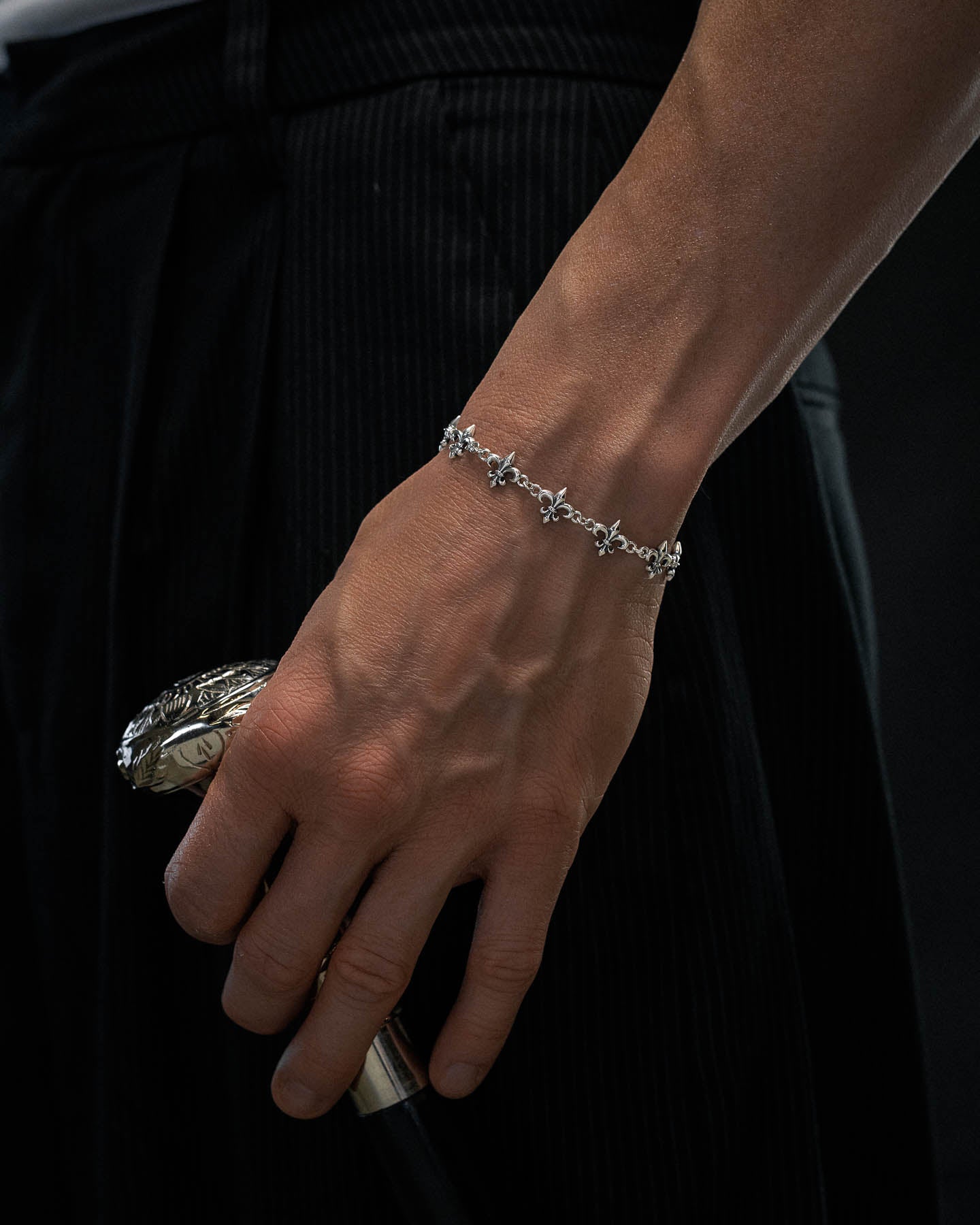 A close-up of a person’s hand holding a cane with a decorative handle, wearing the Statement Collective 925 Sterling Silver "Fleur De Lis" 10mm Bracelet, and dressed in dark pinstripe pants.