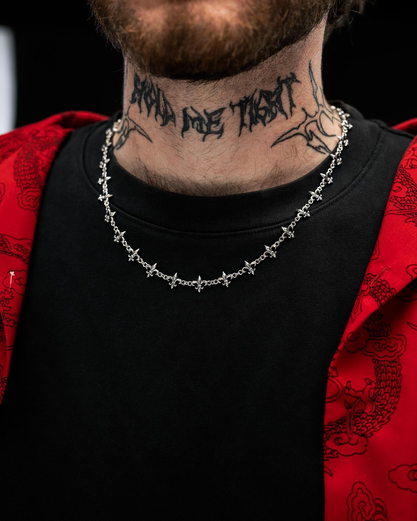 A person wearing a black shirt, a red jacket with a dragon pattern, the Statement Collective 925 Sterling Silver "Fleur De Lis" 10mm Chain, and a neck tattoo that reads AND ONE THAT.