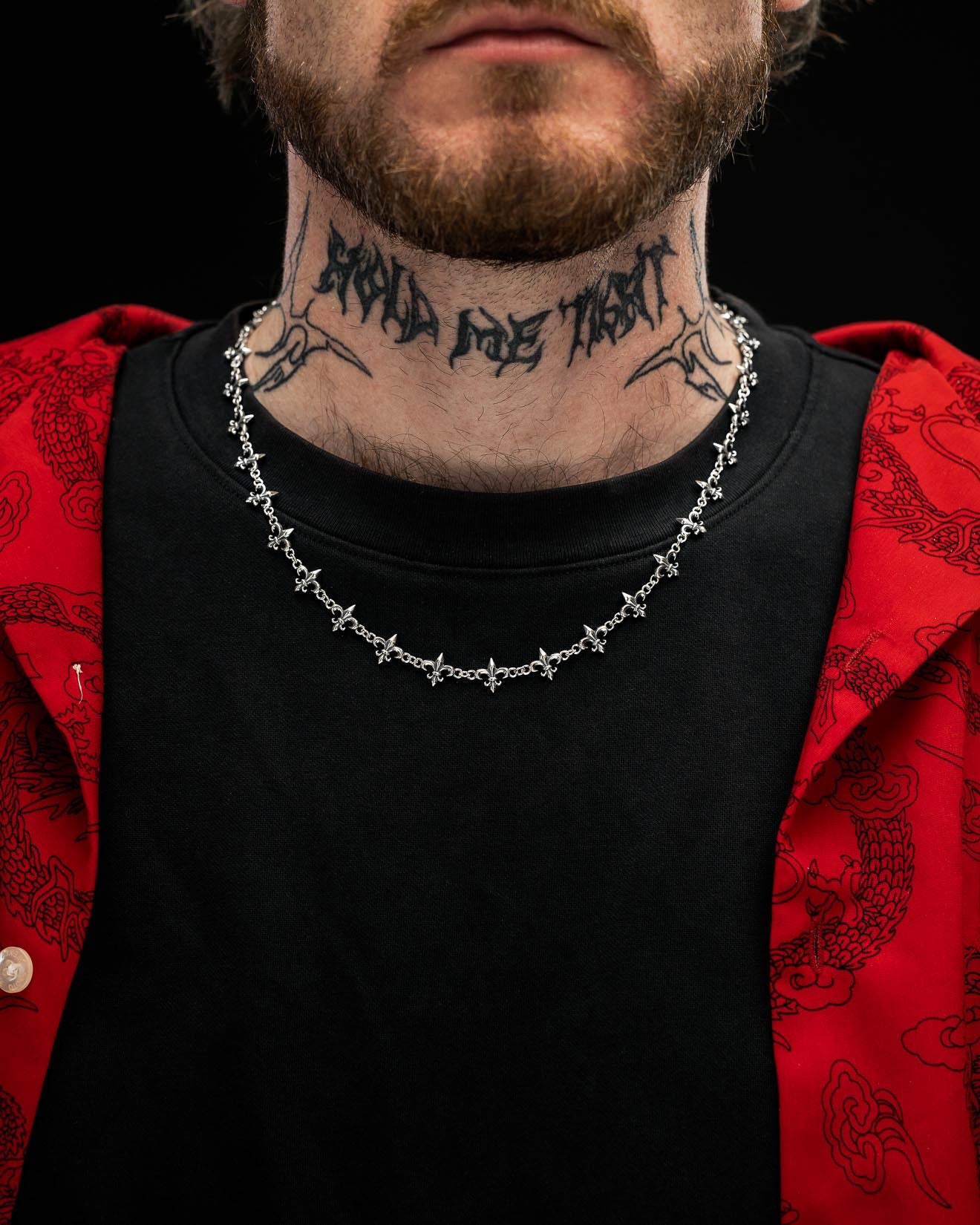 A man with a beard wears a black shirt, a red patterned jacket, a Statement Collective 925 Sterling Silver "Fleur De Lis" 10mm Chain necklace, and has a tattoo reading "Mind My Trust" on his neck.