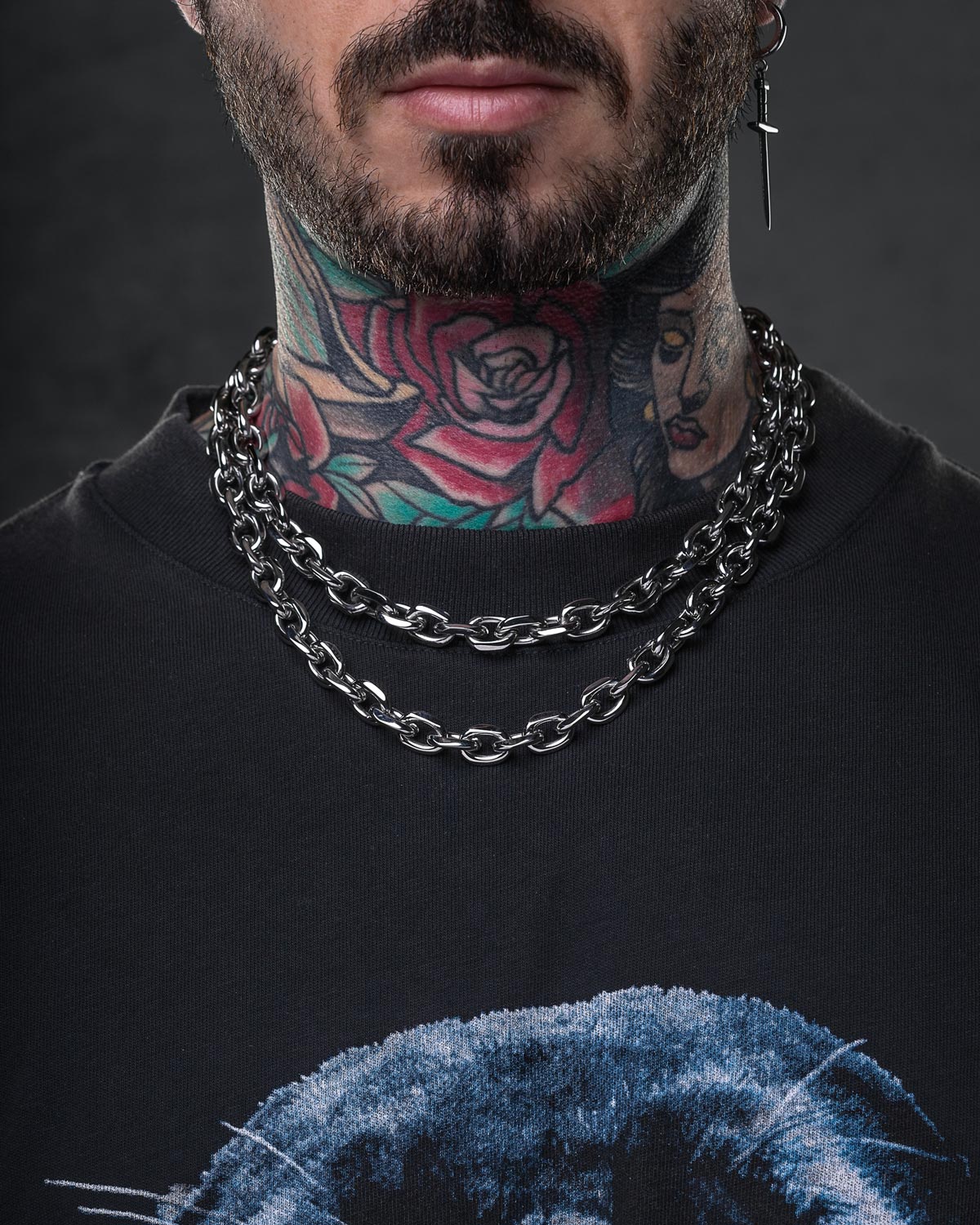 A bearded individual wears the 10mm Cable Chain with layered silver streetwear jewelry, a graphic black shirt, colorful neck tattoos, and a cross-shaped dangling earring.
