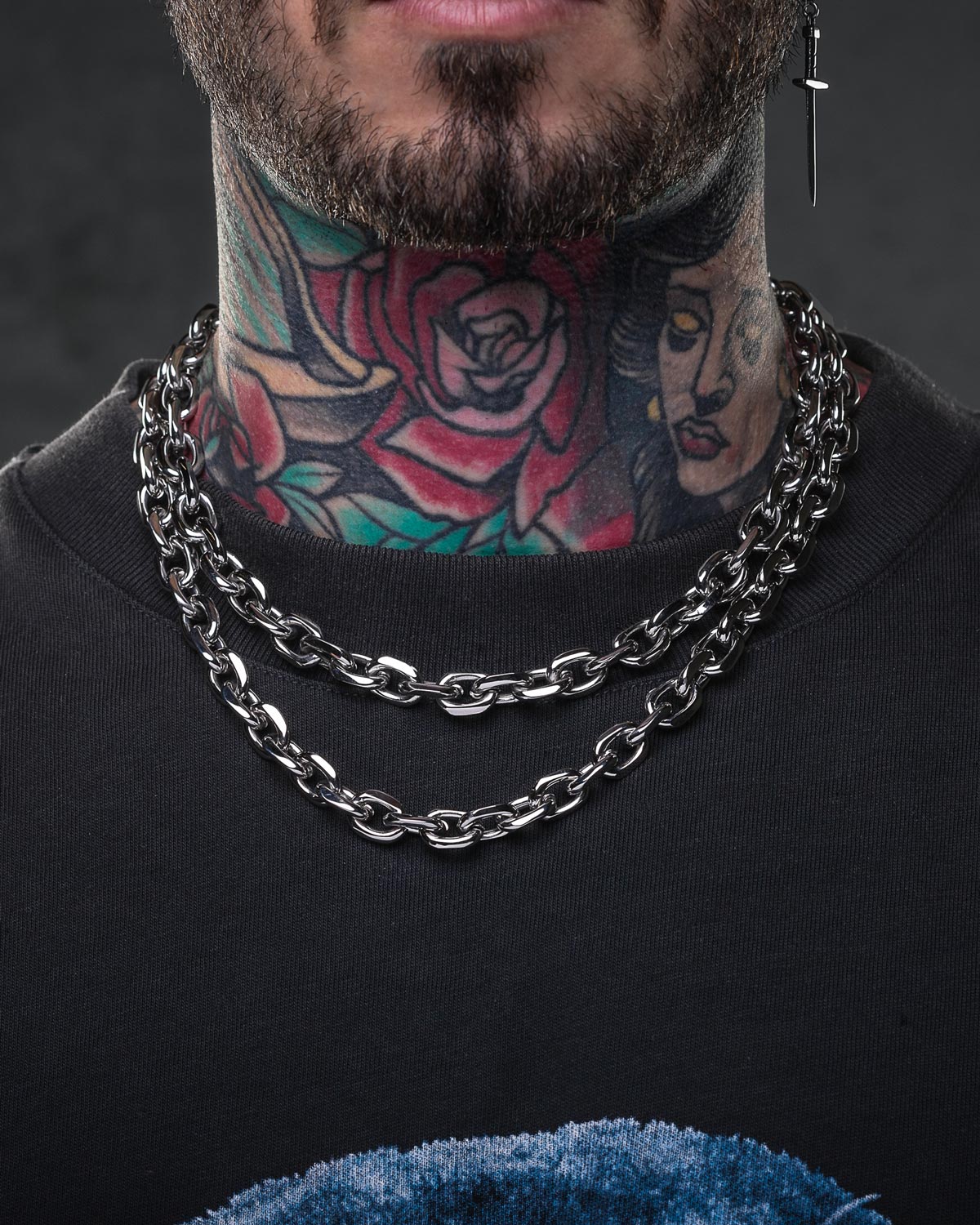 A person with a rose and woman's face tattoo on their neck wears two thick 10mm Cable Chains and a black shirt. Only the lower face and neck are seen. A cross-shaped earring completes the streetwear jewelry look.