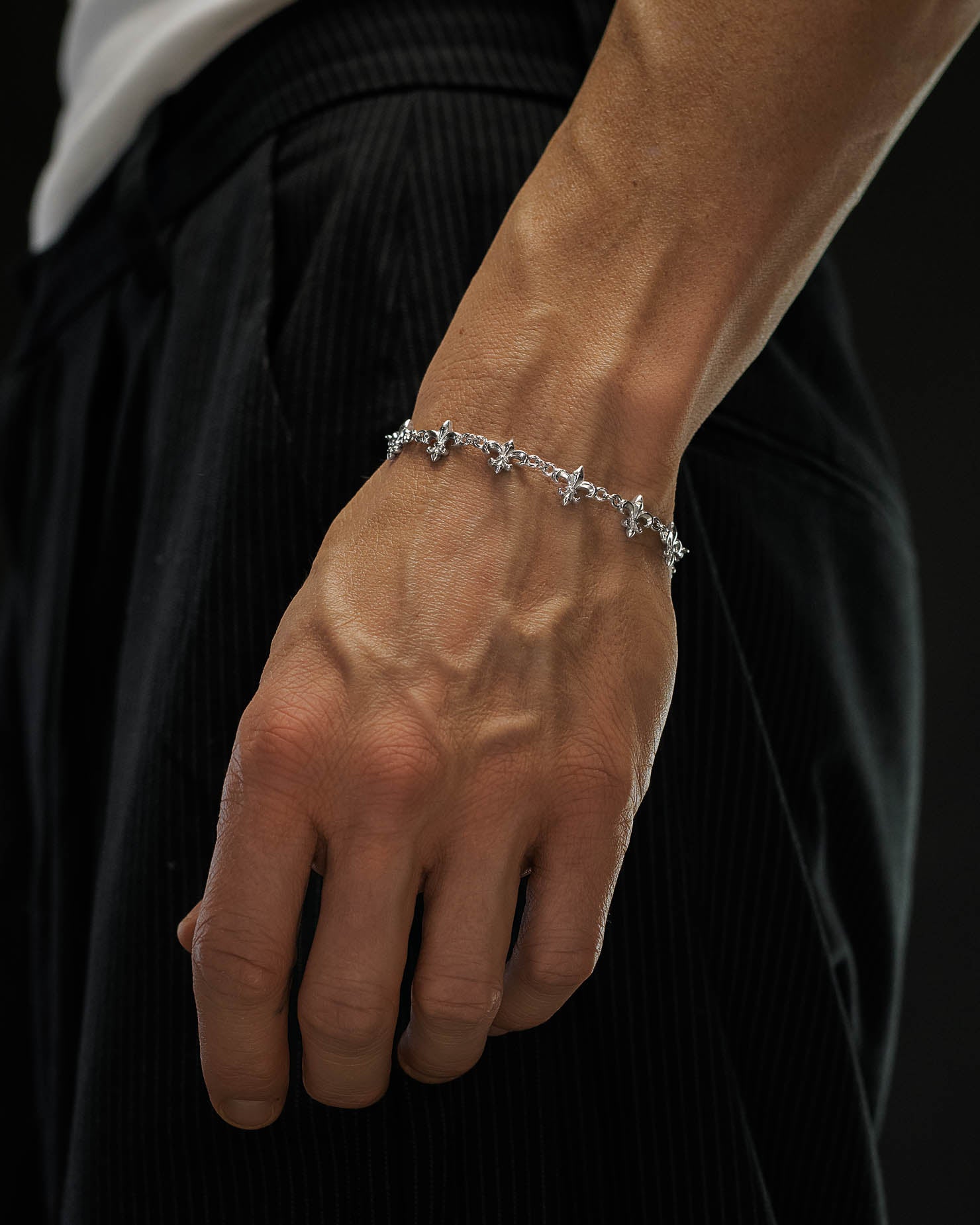 A close-up of a person’s hand wearing the Statement Collective "Fleur De Lis" 10mm Bracelet, shown against black pinstriped pants and a white shirt.