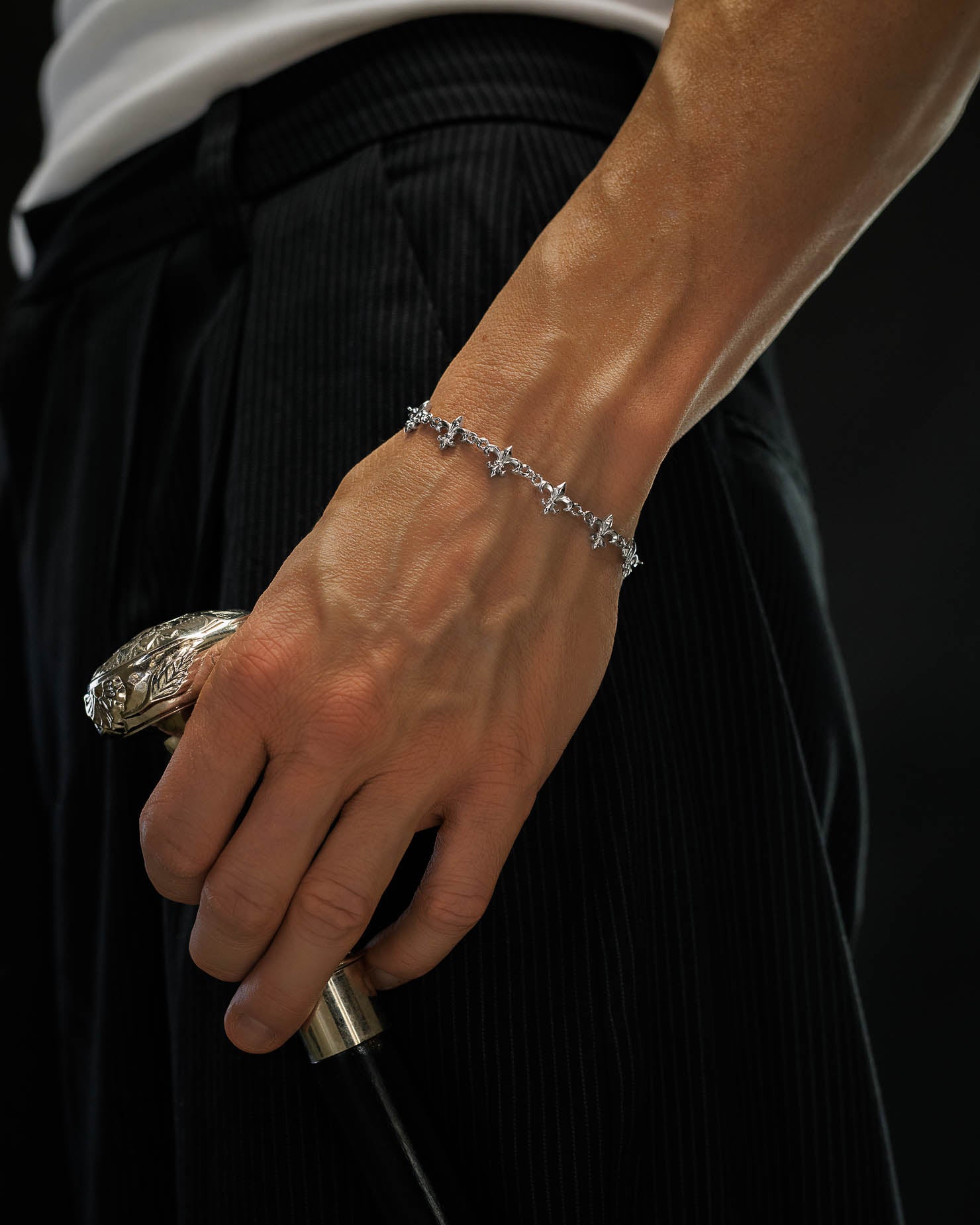 A close-up of a person wearing the Statement Collective "Fleur De Lis" 10mm Bracelet, holding an ornate cane. The person is dressed in black pinstripe pants and a white shirt, set against a black background.