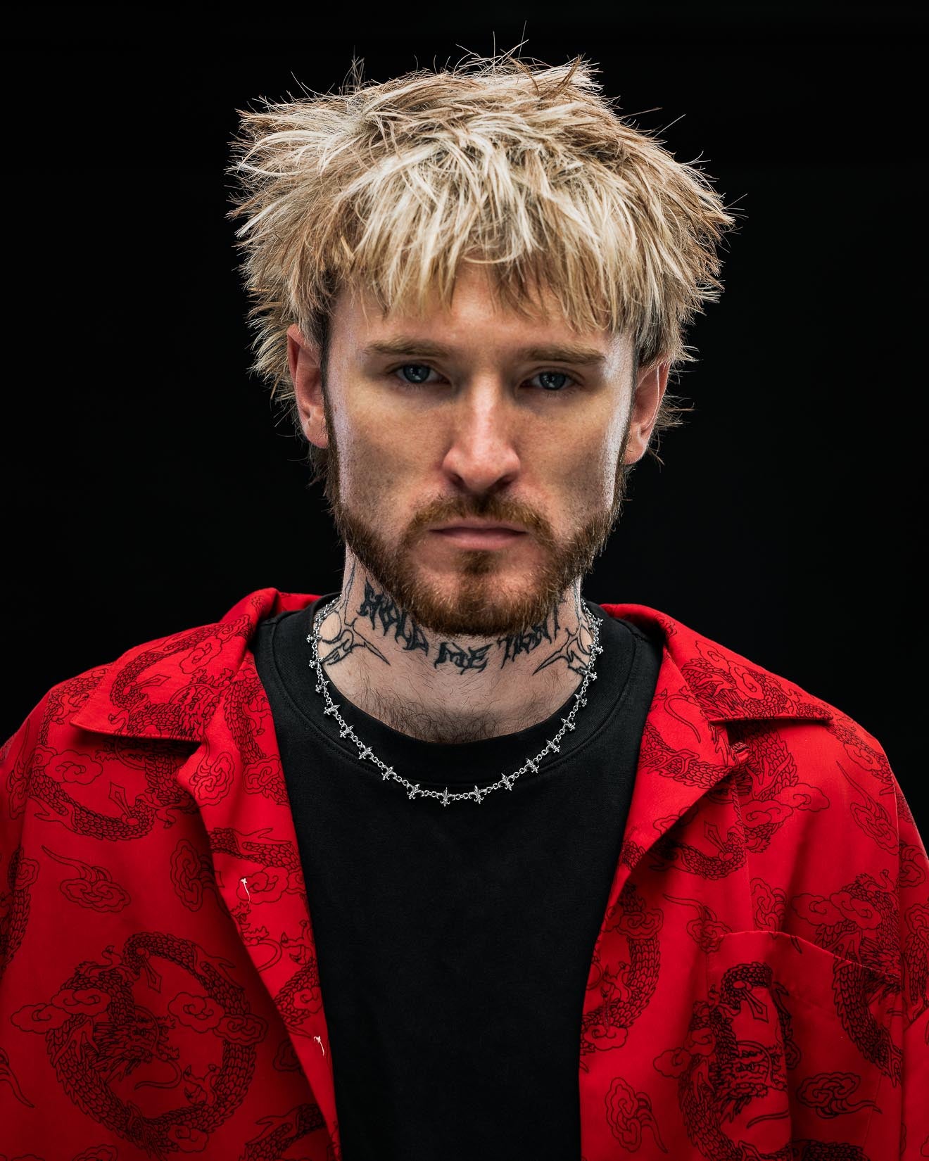 A person with shaggy blond hair, a full beard, and neck tattoos wears Statement Collective’s "Fleur De Lis" 10mm Chain along with a red patterned shirt layered over a black top, staring seriously at the camera against a dark background.