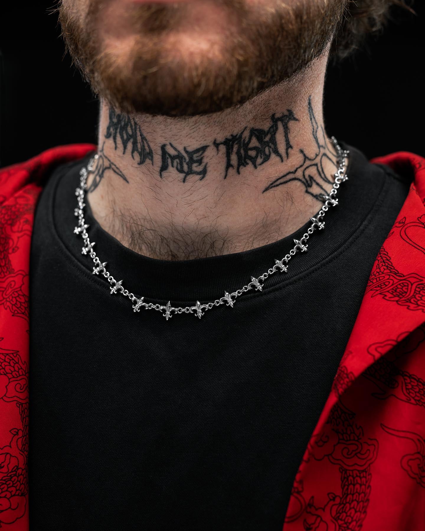 A close-up of a person wearing the Statement Collective "Fleur De Lis" 10mm Chain necklace, paired with a black shirt and a patterned red jacket. Only the lower face and neck—featuring a stylized text tattoo—are visible.
