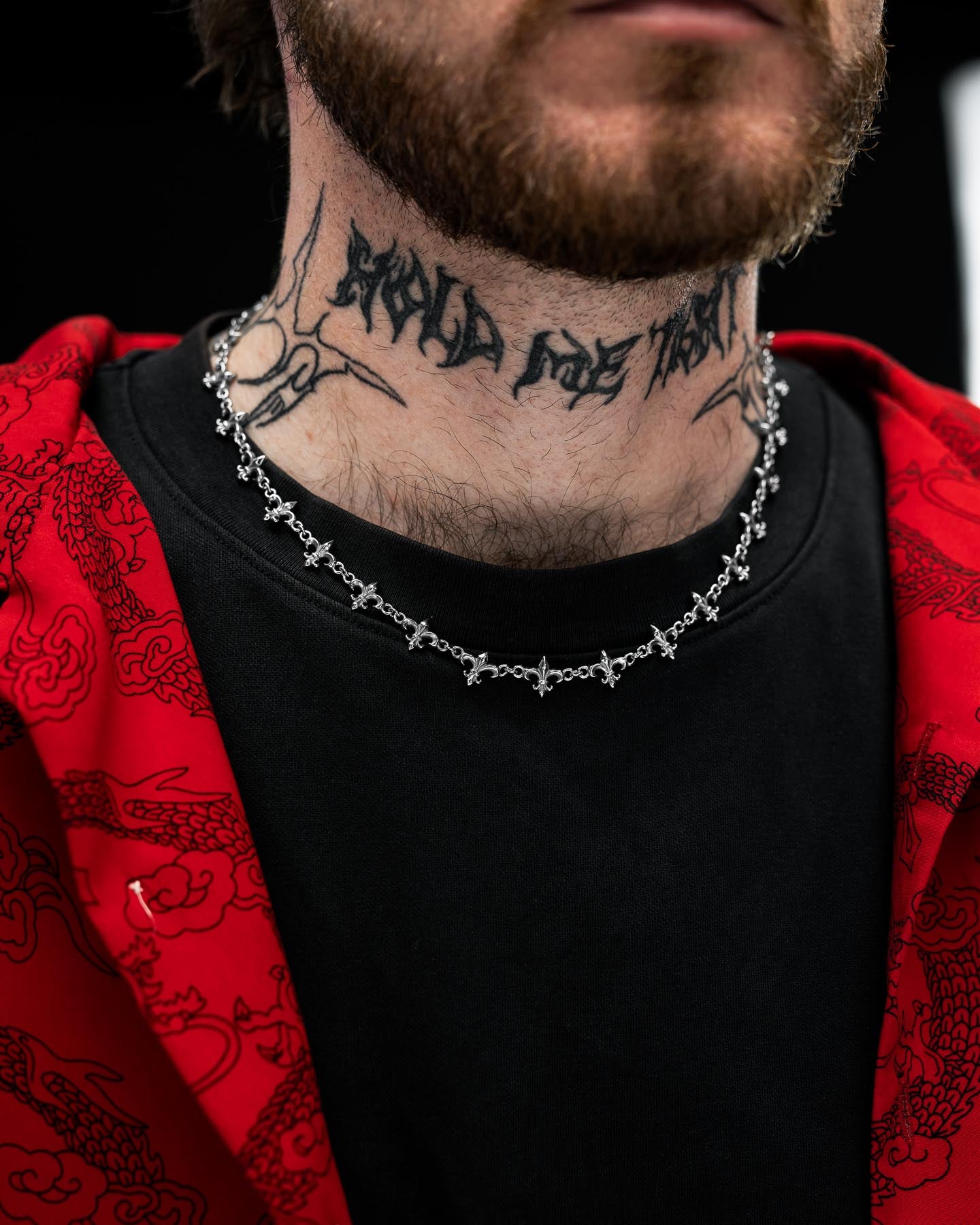 A close-up of a man's neck adorned with the Statement Collective "Fleur De Lis" 10mm Chain. He wears a black shirt and a red patterned over-shirt, with only the lower half of his face visible, showcasing his tattoo and silver chain necklace.