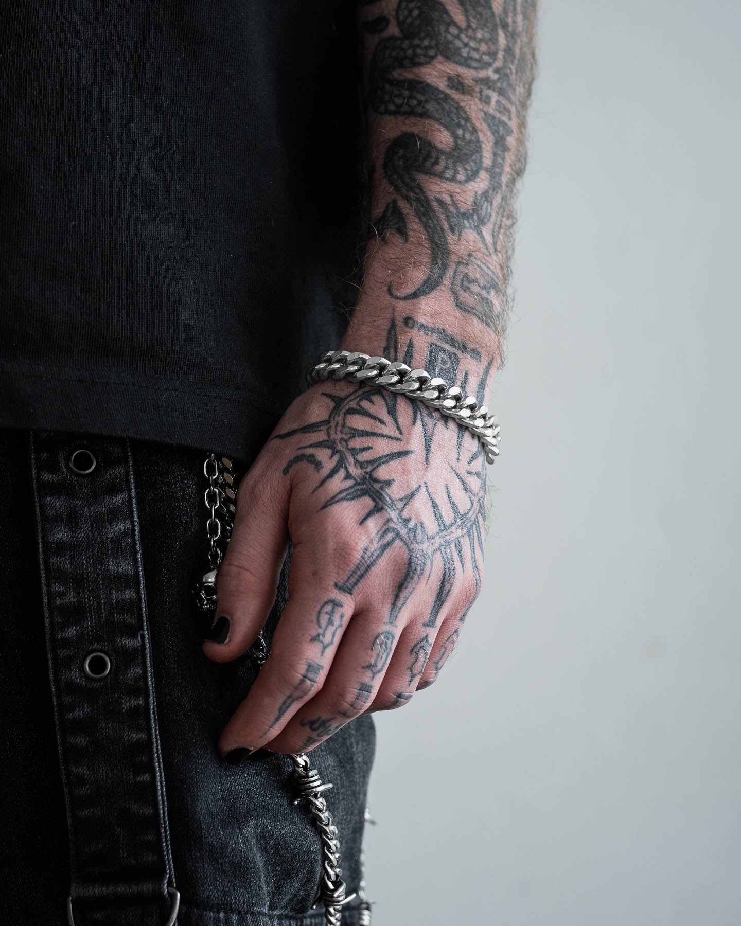 A close-up of a tattooed arm and hand wearing the 11mm Cuban Bracelet. The person, dressed in a black shirt and black denim pants with metal chains, displays bold tattoos for an edgy look.