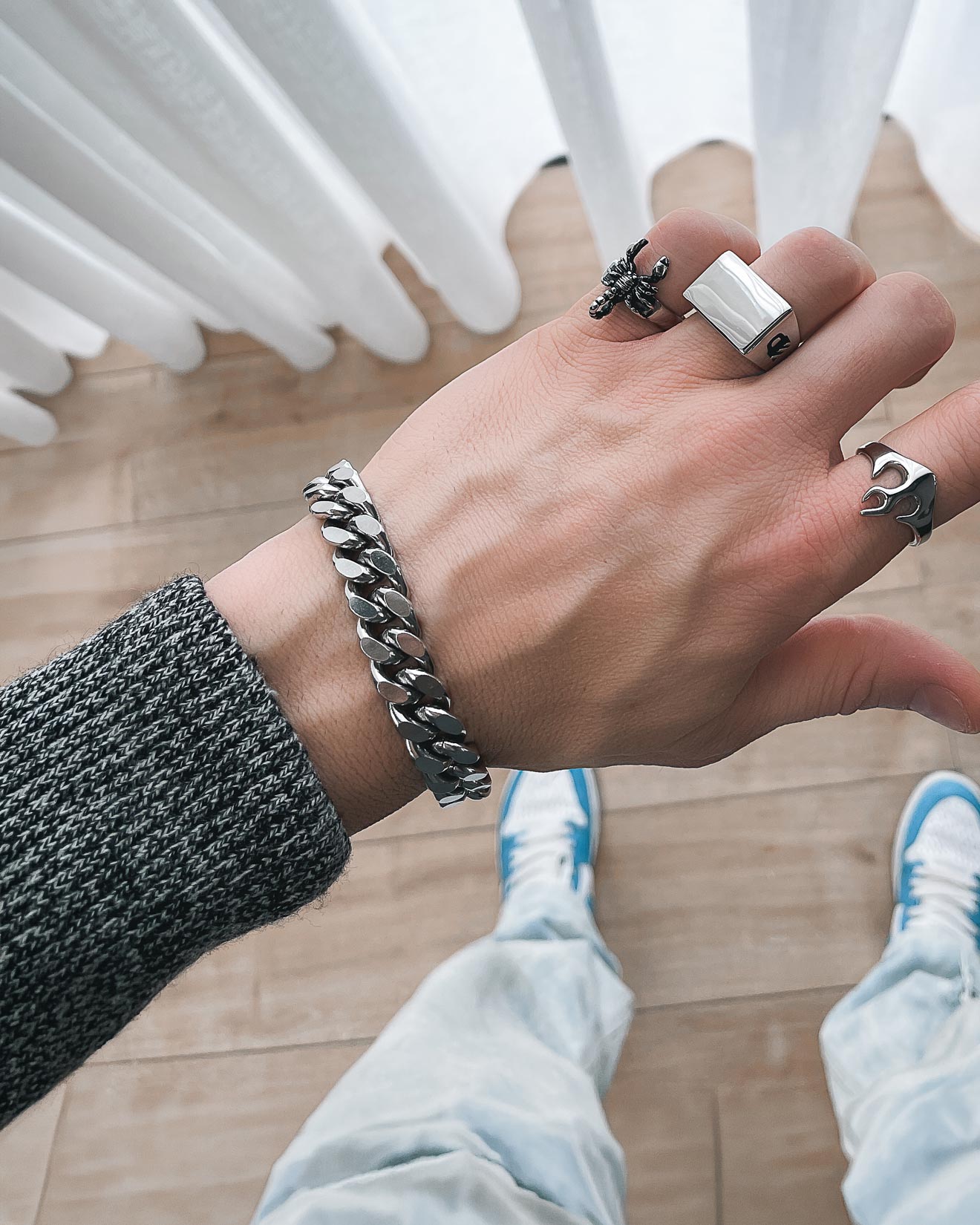A person wearing the 11mm Cuban Bracelet by Statement Collective and several rings extends their hand over a wooden floor, dressed in a textured gray sweater, light blue jeans, and blue-and-white sneakers with white curtains in the background.