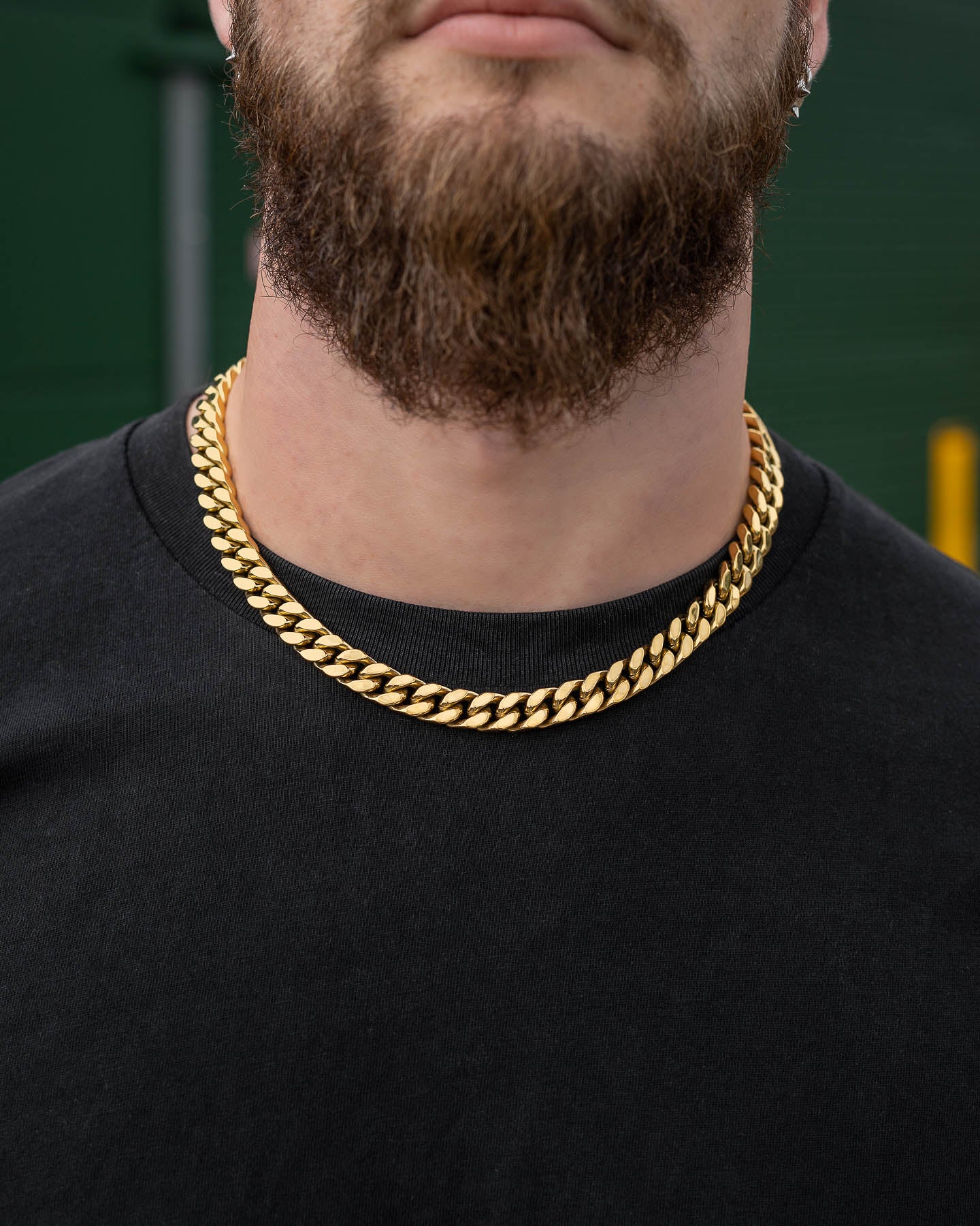 A close-up of a person with a brown beard wearing a black shirt and two thick 11mm Cuban Chain (Gold) necklaces, standing outdoors with a blurred background.
