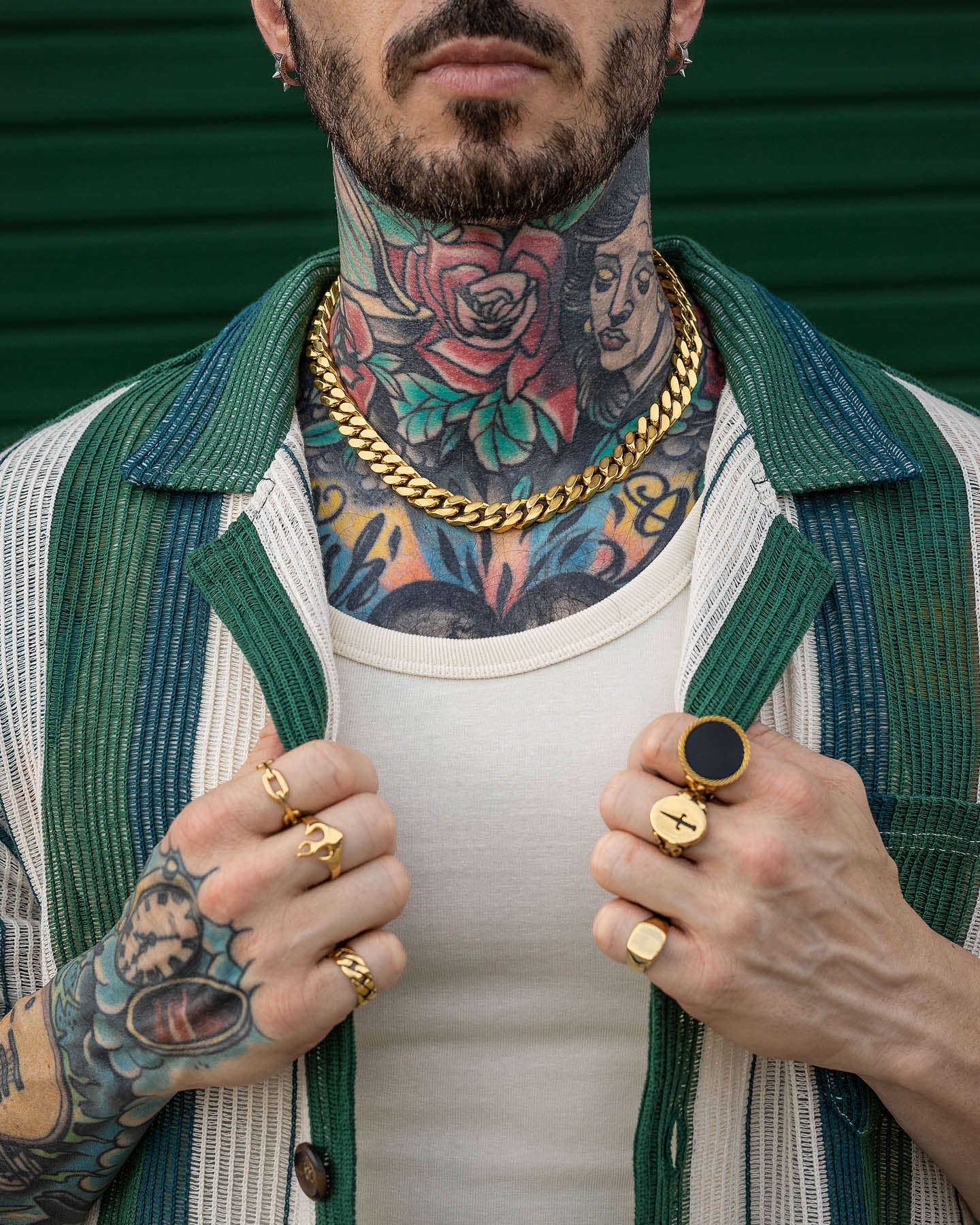 A tattooed man wearing gold rings, an 11mm Cuban Chain (Gold) necklace that's non-tarnish and waterproof, and a green-and-white striped shirt over a white tank top, showcasing tattoos on his neck, hands, and chest.