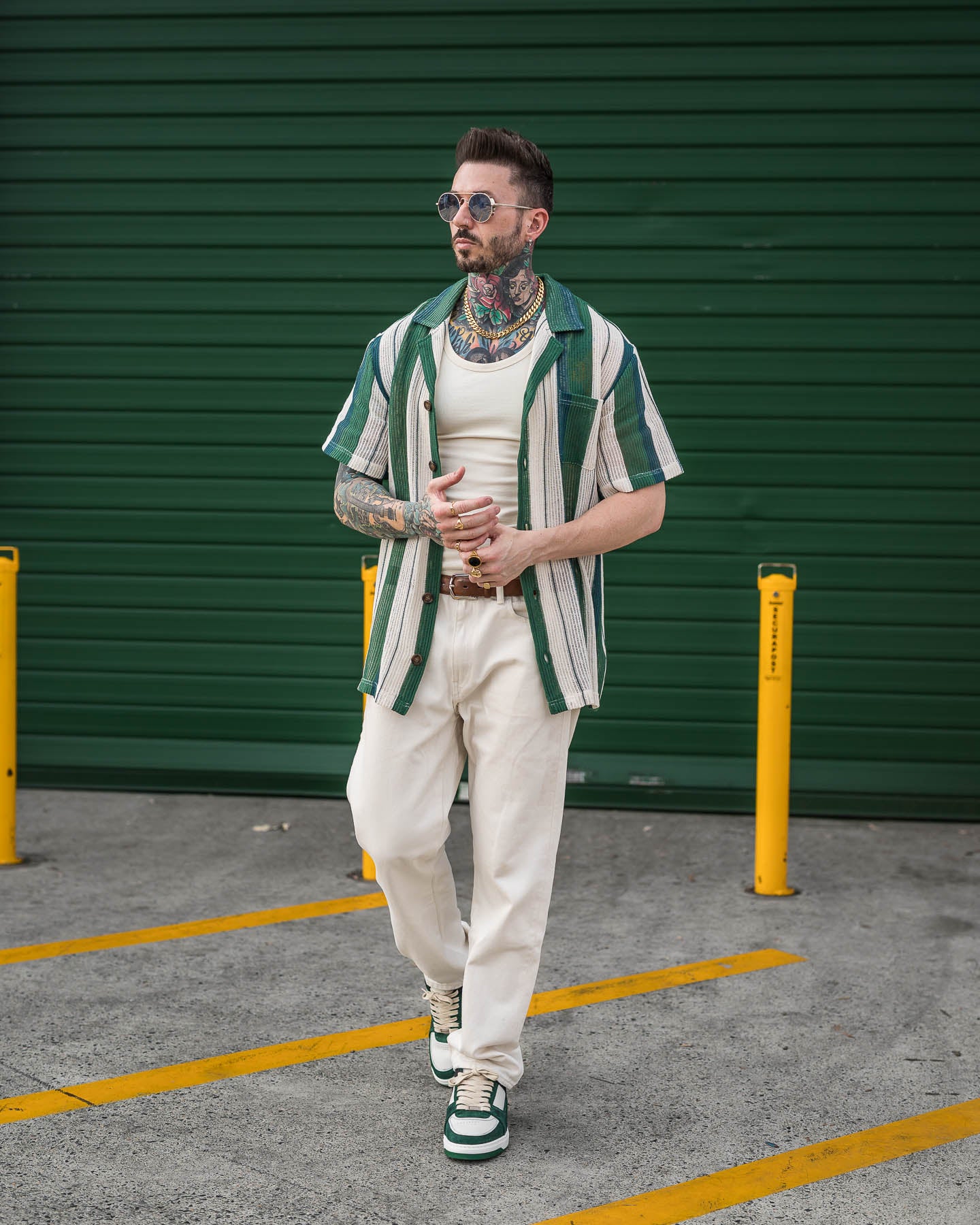 A man with tattoos, sunglasses, and styled hair stands in front of a green garage door, wearing a green and white striped shirt, white pants, matching sneakers, and the 11mm Cuban Chain (Gold). Yellow poles and lines are visible.