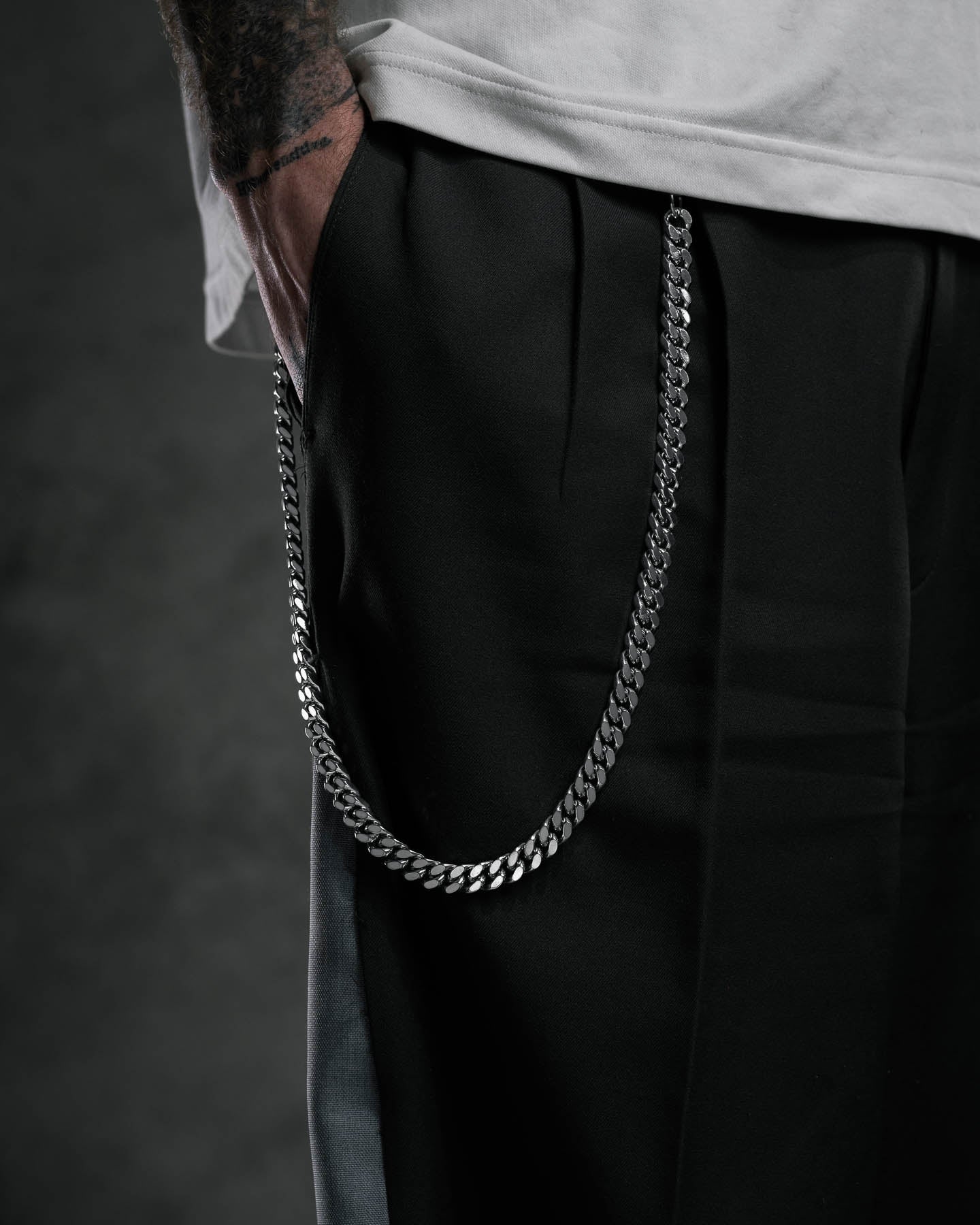 A person wears black pants with an 11mm Cuban Wallet Chain in 316L stainless steel attached to the pocket and a white shirt, showing only the lower torso and one tattooed arm with a hand in the pocket against a dark background.