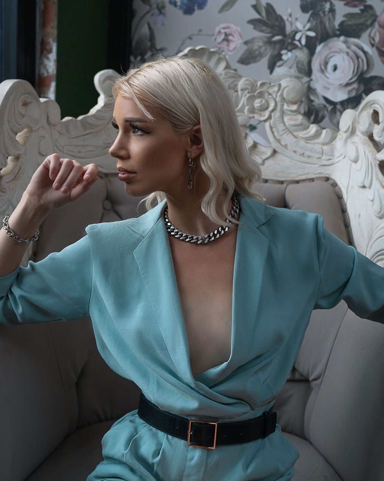 A platinum blonde woman in a light blue suit and the 13mm Chunky Cuban Chain sits pensively on an ornate cream chair, gazing to the side. Floral wallpaper is visible in the background.