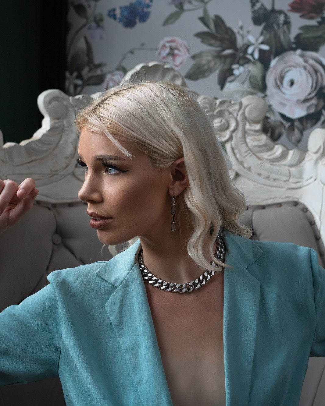 A platinum blonde woman in loose waves wears a pale blue blazer and the 13mm Chunky Cuban Chain choker, seated on an ornate tufted chair against floral wallpaper, gazing thoughtfully to the side.