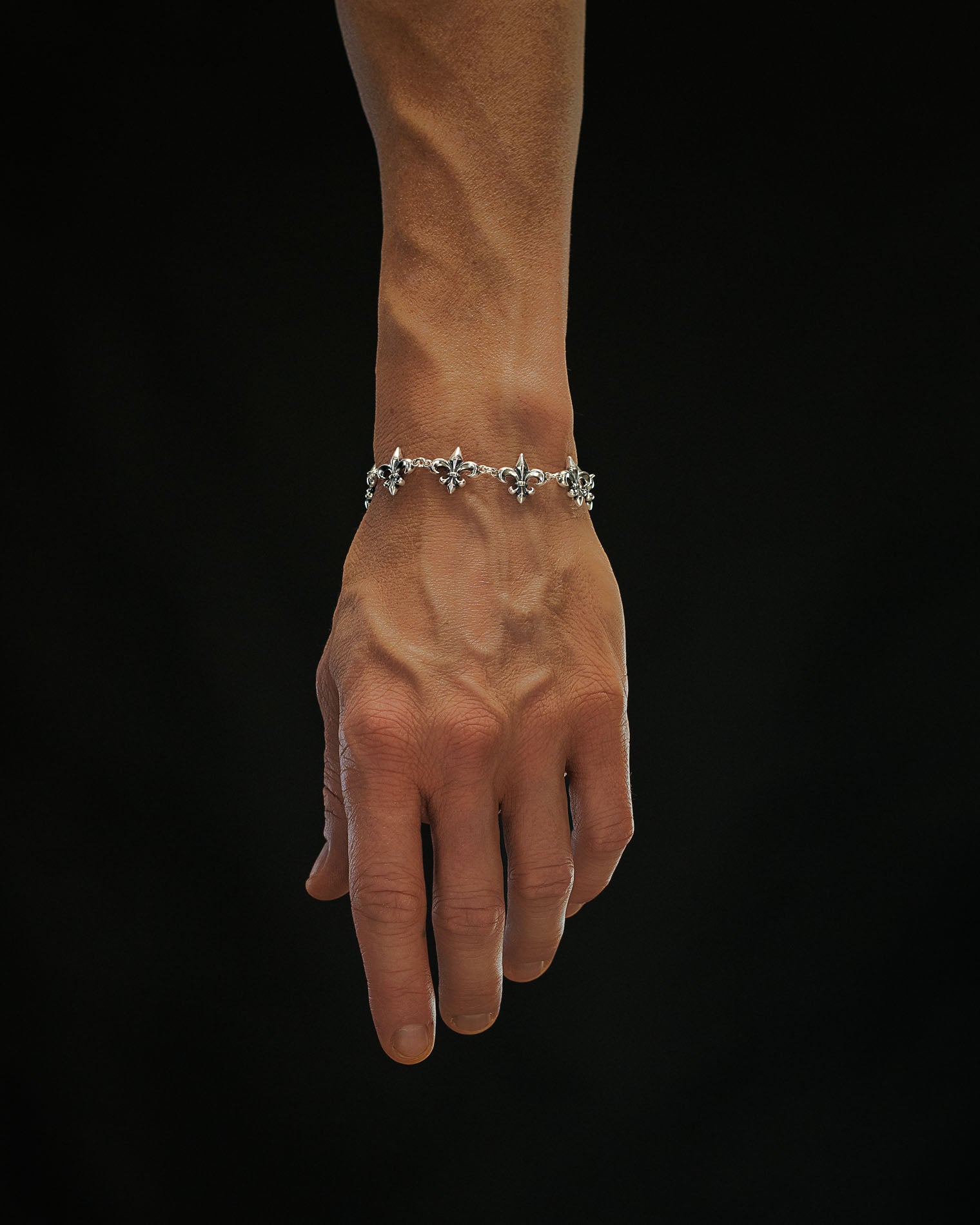 A hand with visible veins wears the Statement Collective 925 Sterling Silver "Fleur De Lis" 15mm Bracelet, displayed against a solid black background.