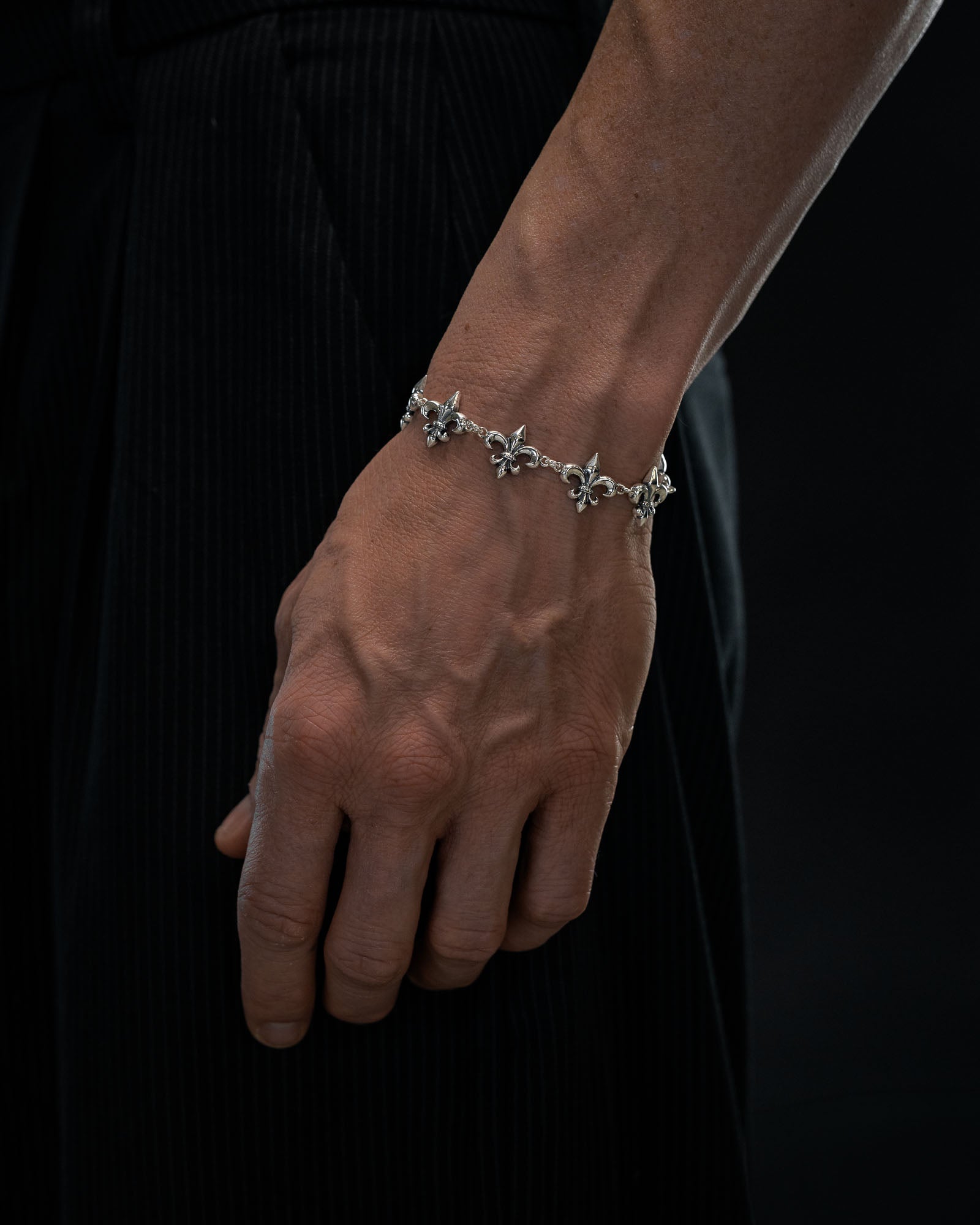 A close-up of a person's left hand wearing the Statement Collective 925 Sterling Silver "Fleur De Lis" 10mm Bracelet, featuring delicate flower-inspired charms, against a dark background. The person is dressed in black pinstriped fabric.