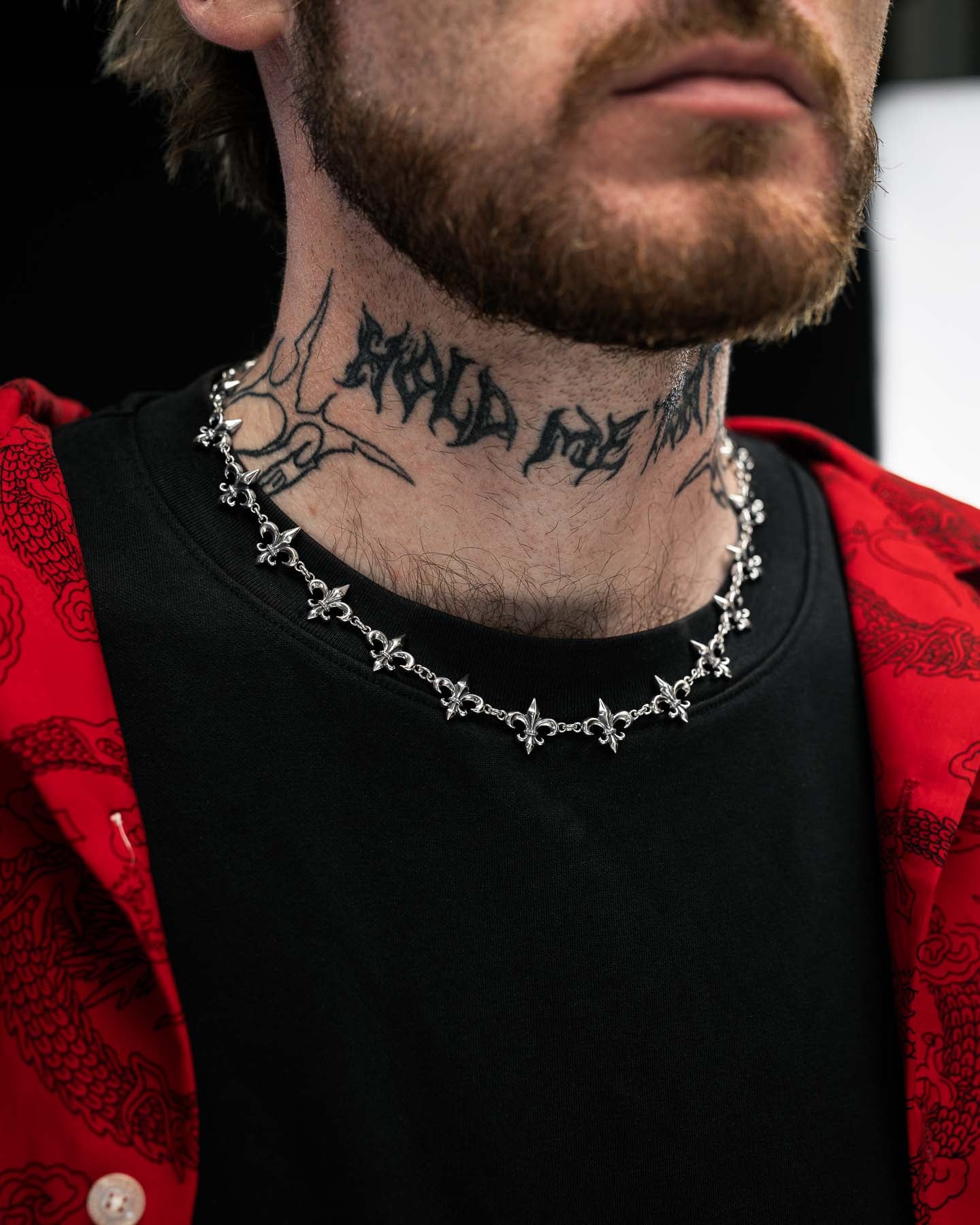 A person with a beard and neck tattoo wears the Statement Collective 925 Sterling Silver "Fleur De Lis" 15mm Chain, paired with a black shirt and a red dragon-patterned jacket.