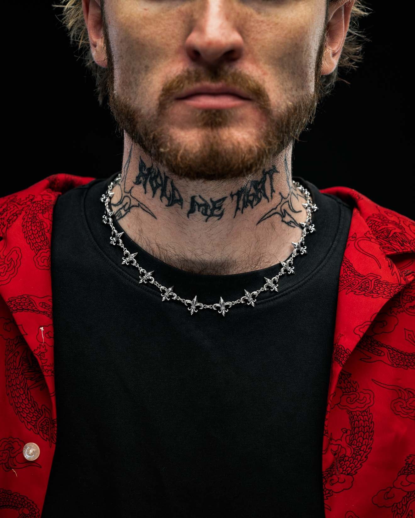 A man with a beard wears a red jacket, black shirt, a Statement Collective 925 Sterling Silver "Fleur De Lis" 15mm Chain, and has a neck tattoo that reads HOLD ME TIGHT. The image focuses on his lower face, neck, and chest against a black background.