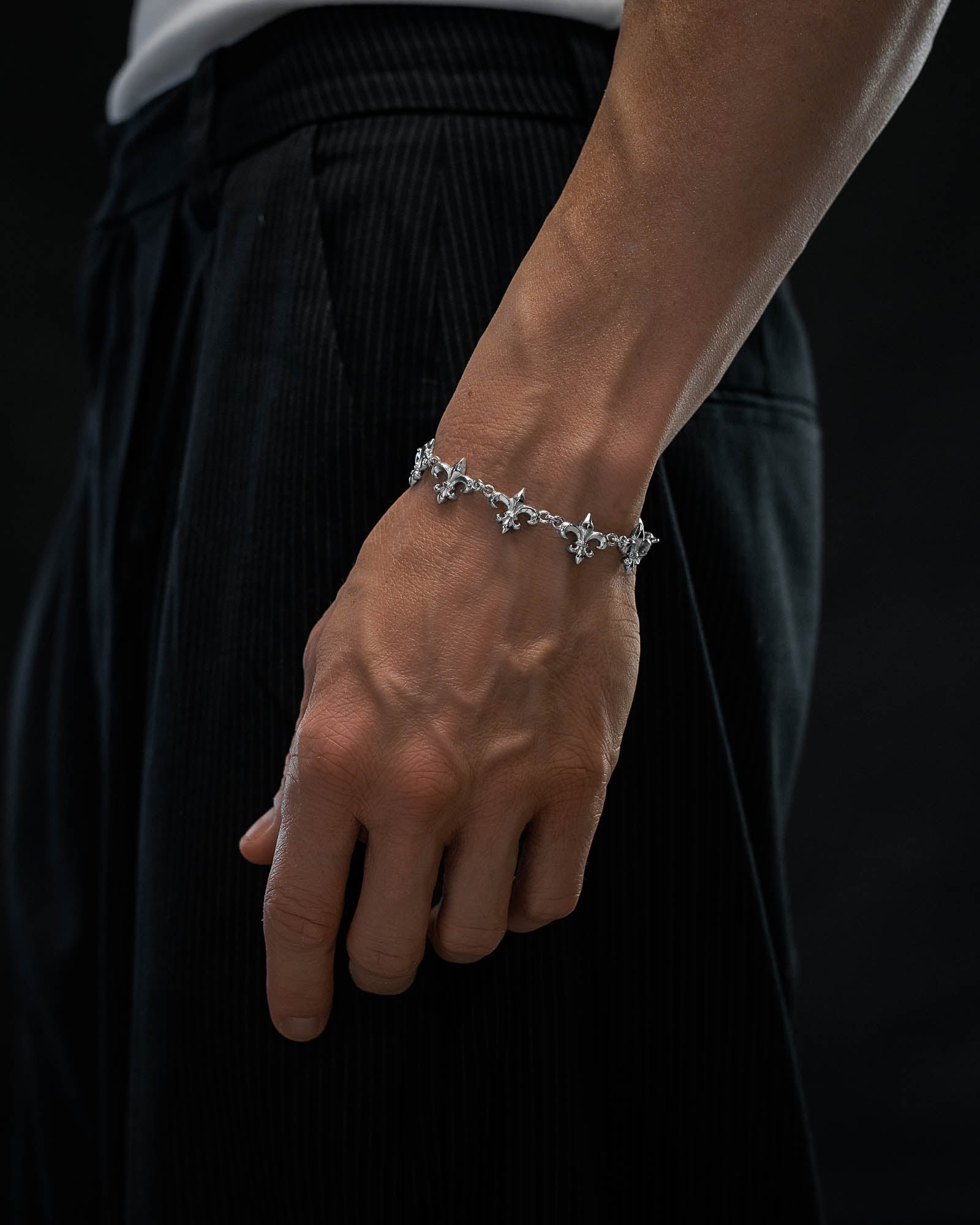 A close-up of a person's hand wearing the "Fleur De Lis" 15mm Bracelet by Statement Collective, featuring intricate silver star-shaped links. The person is dressed in black pinstriped pants and a white shirt, set against a dark background.