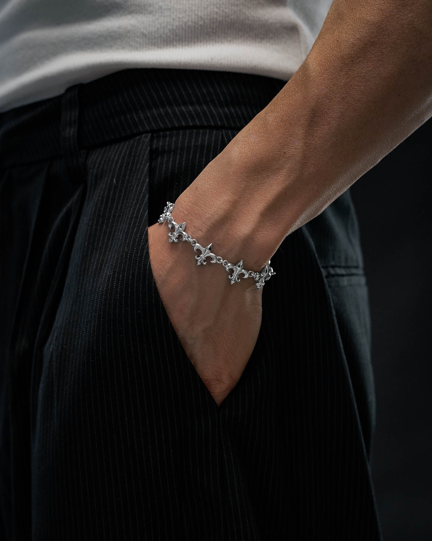 A person dressed in dark pinstripe pants and a white shirt has their hand in their pocket, showcasing the Statement Collective "Fleur De Lis" 15mm Bracelet on their wrist.