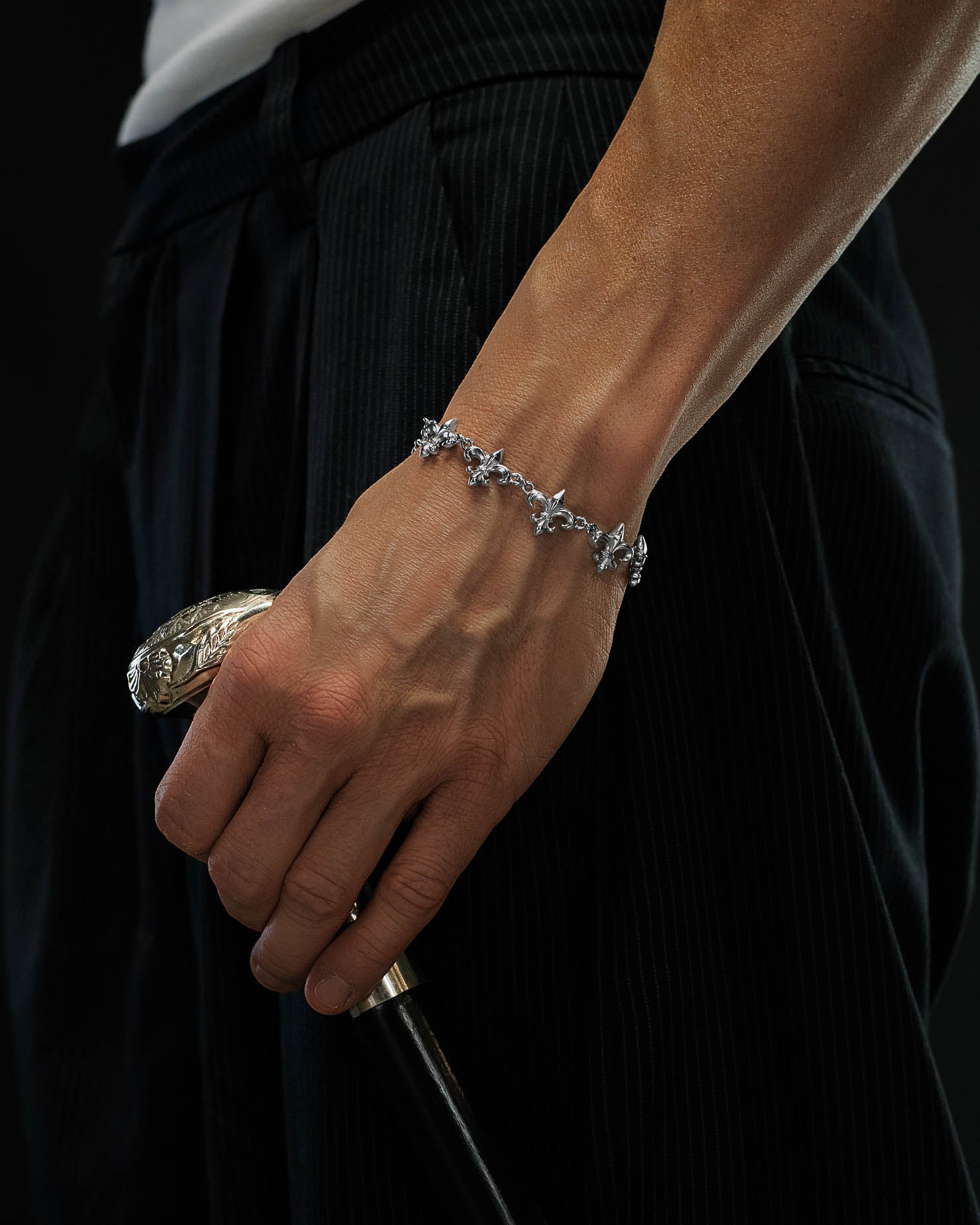 Wearing Statement Collective’s "Fleur De Lis" 15mm silver bracelet, a person holds a cane with an ornate handle, dressed in dark pinstripe pants and a white shirt, set against a black background.