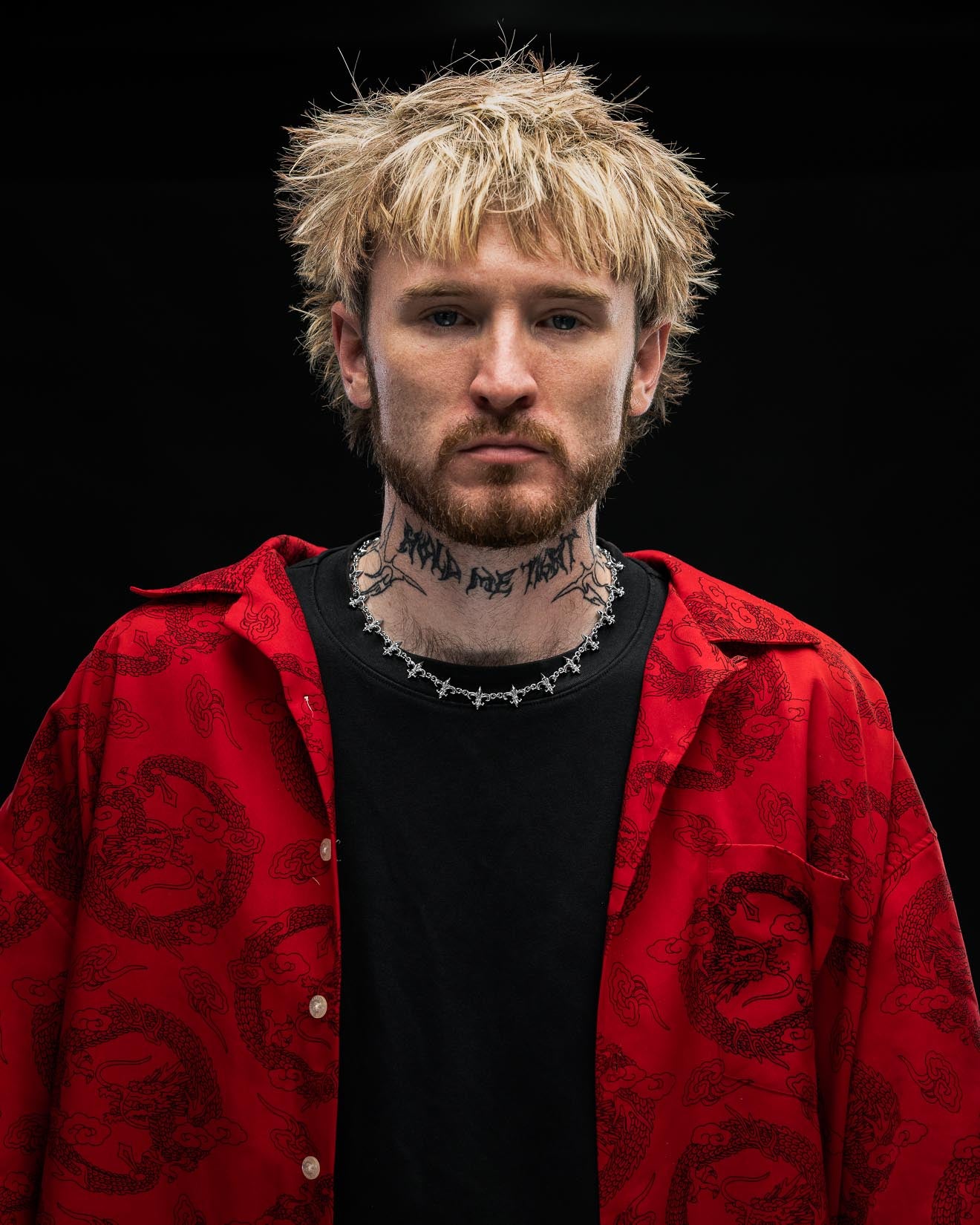 A man with messy blond hair, a beard, and a neck tattoo wears a black shirt, a red patterned jacket, and the "Fleur De Lis" 15mm Chain from Statement Collective, posing against a dark background.