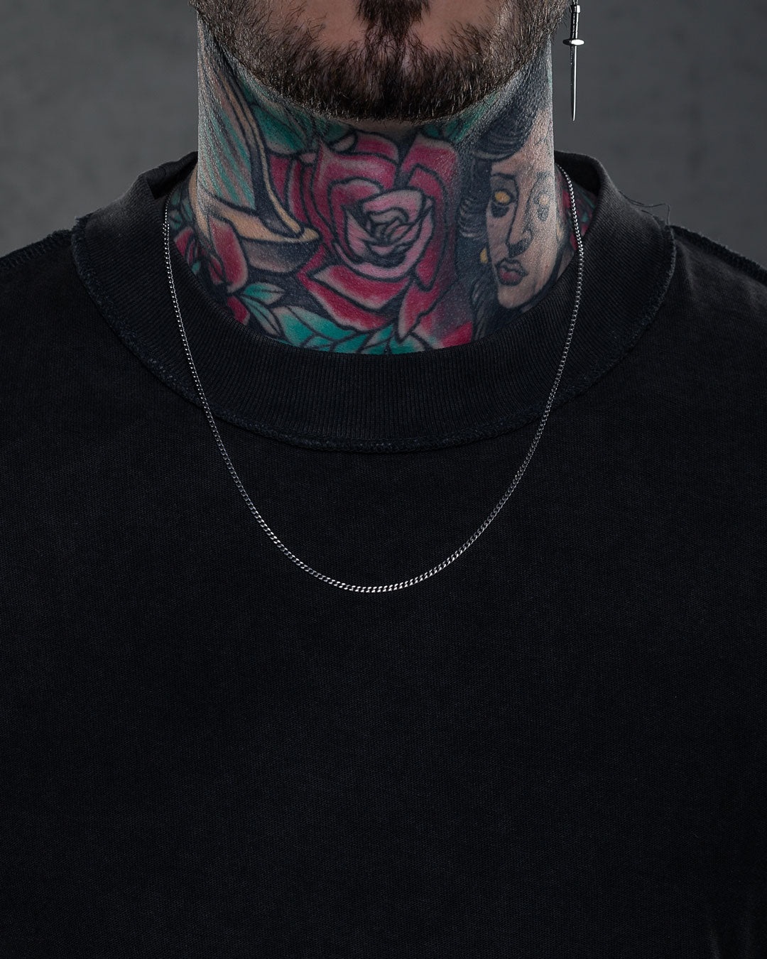 A person wearing a plain black shirt, a 2.3mm Curb Chain necklace, and a cross earring in their left ear. Their neck is covered in colorful tattoos of roses and a woman's face.